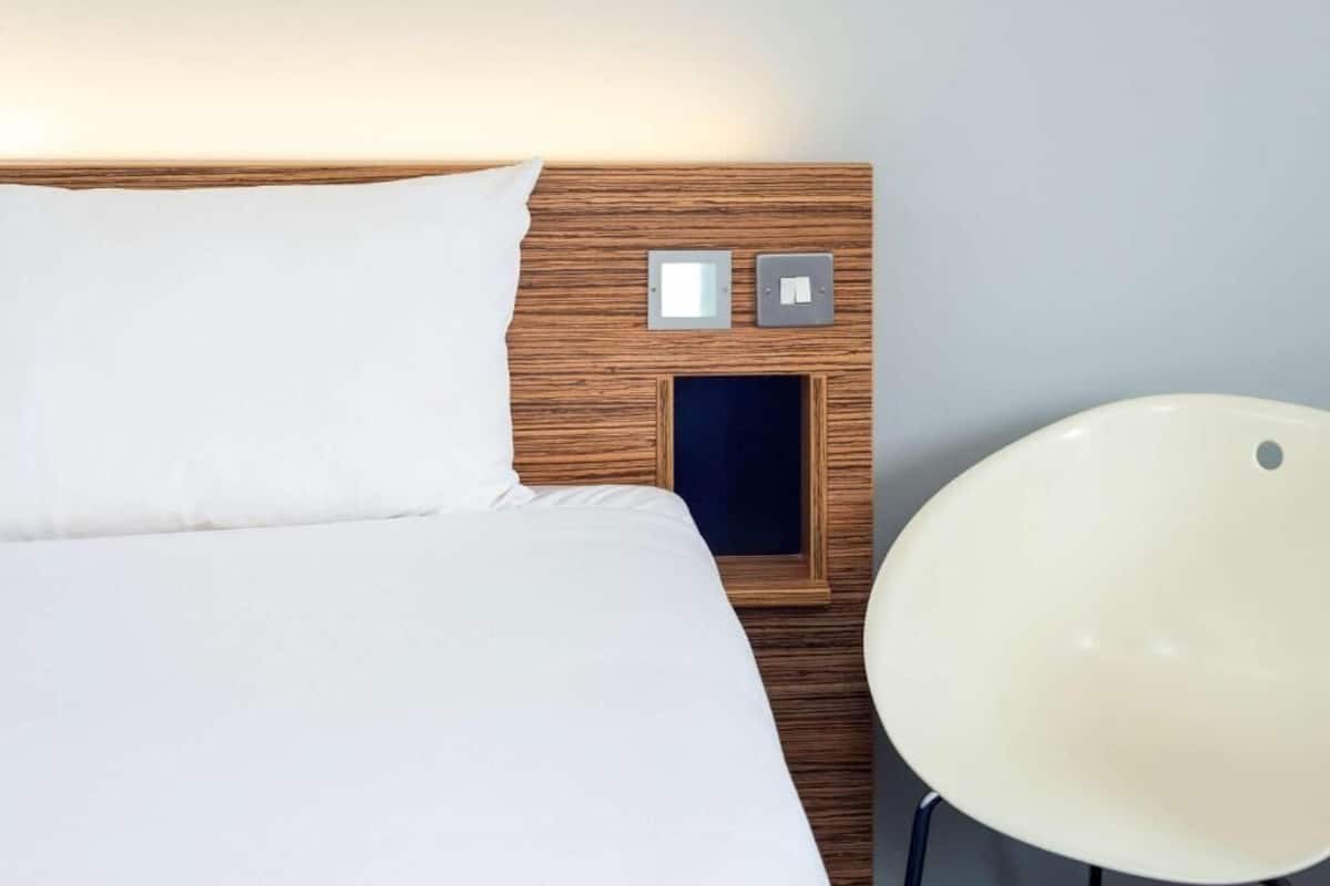 A neatly made bed with white linens, a wooden headboard, and a bedside table with two switches and a small drawer.