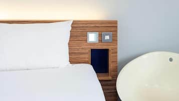 Un lit bien fait, avec du linge de lit blanc, une tête de lit en bois et une table de chevet dotée de deux interrupteurs et d’un petit tiroir.