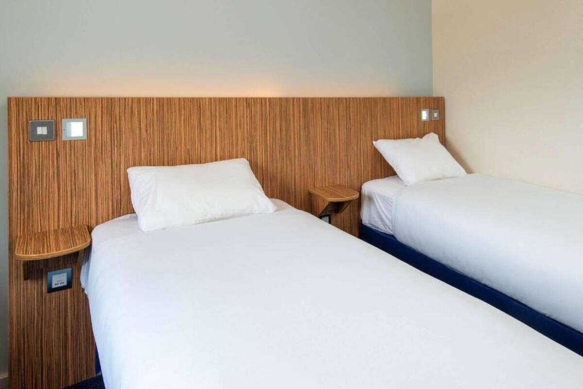 A hotel room with two single beds, wooden headboard, bedside tables, and wall-mounted light switches.