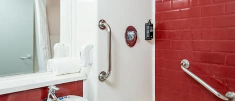 Une salle de bain avec un lavabo blanc, une baignoire et des murs recouverts de carreaux rouges. Des barres d'appui sont installées près de la baignoire et un sèche-mains est fixé au mur.