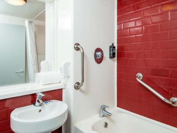 Un baño con un lavamanos blanco, una bañera y paredes cubiertas de baldosas rojas. Se observan barras de apoyo cerca de la bañera y un secamanos en la pared.