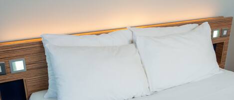 A neatly made bed with white linens and pillows, a wooden headboard with built-in lighting, and a small bedside table with a lamp.