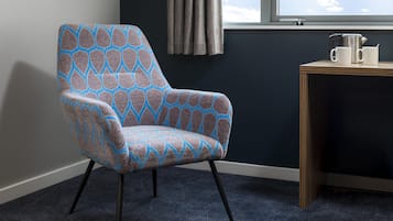 A room with a patterned armchair, a small table with a kettle and cups, a window with curtains, and a dark wall.