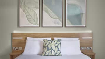 A neatly made bed with white linens and a leaf-patterned pillow, flanked by wooden nightstands with electrical outlets. Above, three framed artworks with abstract leaf designs are hung on the wall.