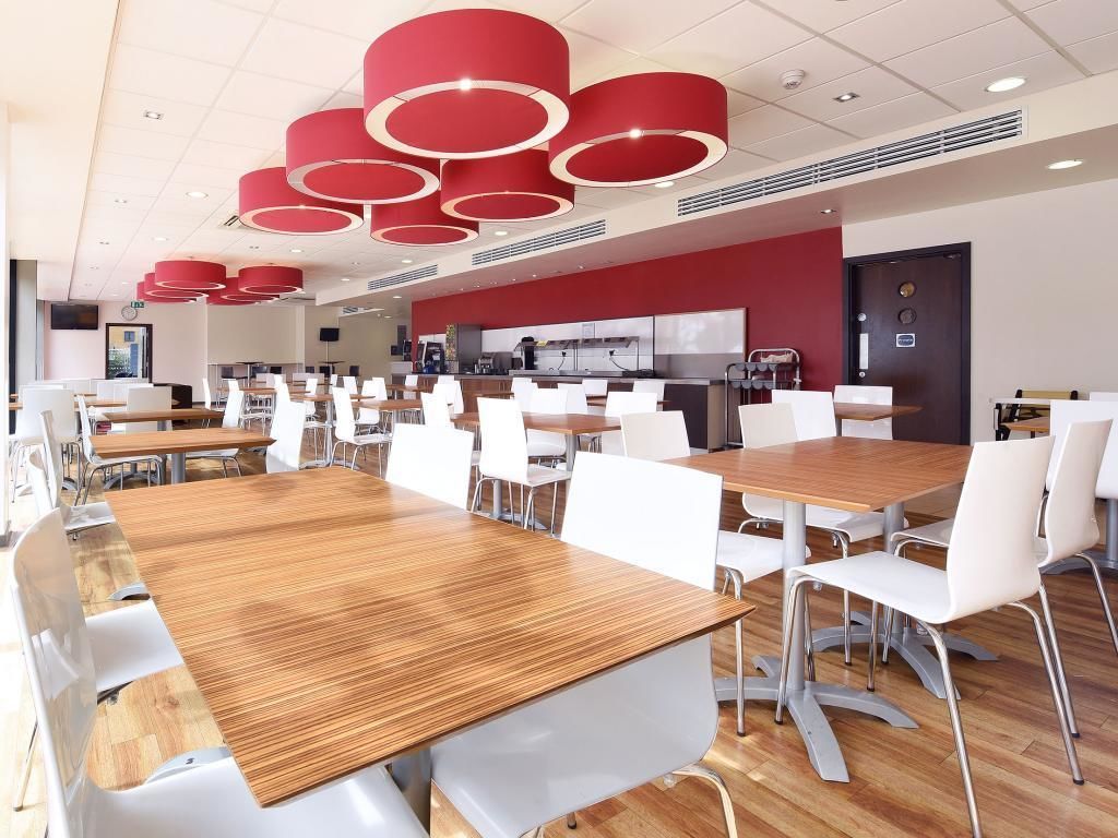 A modern dining area with multiple tables and chairs, a kitchen in the background, and red circular ceiling lights.