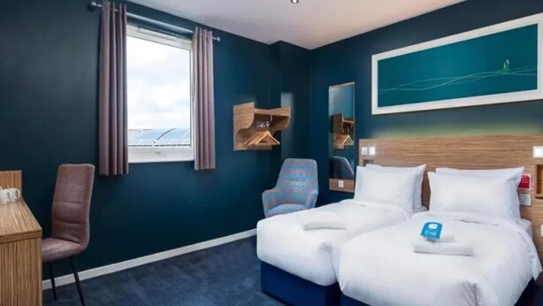Ein Hotelzimmer mit einem großen Bett, einem Schreibtisch, einem Sessel, einem Fenster mit Vorhängen, einem Spiegel und einem Bild an der Wand.