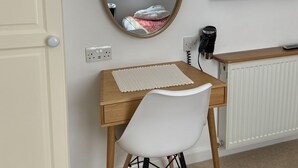 Un dormitorio con un escritorio de madera, una silla blanca, un espejo redondo, una cama con una manta estampada en rojo y blanco, un televisor y un radiador.