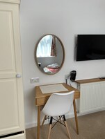 Un dormitorio con un escritorio de madera, una silla blanca, un espejo redondo, una cama con una manta estampada en rojo y blanco, un televisor y un radiador.