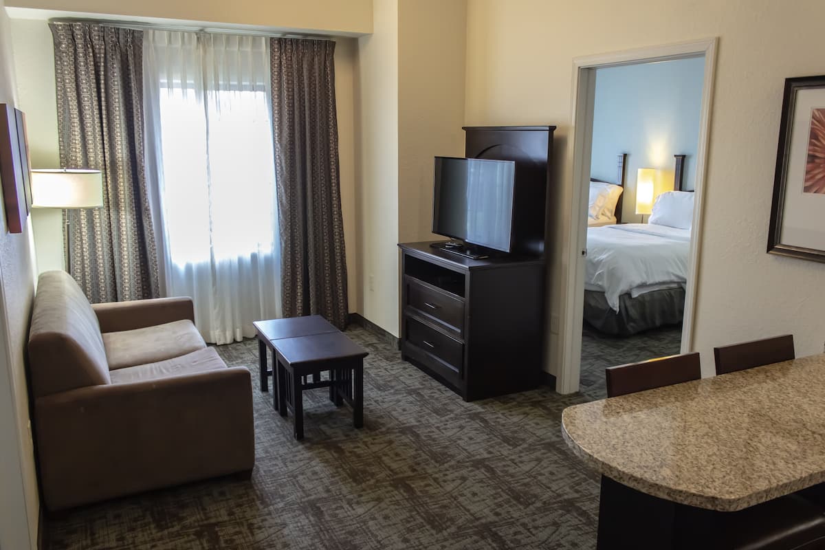 Habitación de hotel con sofá, televisor sobre un mueble bajo, una mesita pequeña, una mesa de comedor con sillas y una cama con mesita de noche.