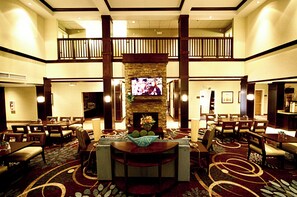 A hotel lobby with a central seating area, a television mounted on a stone wall, and a balcony overlooking the lobby. - Staybridge Suites Minot by IHG (Minot)