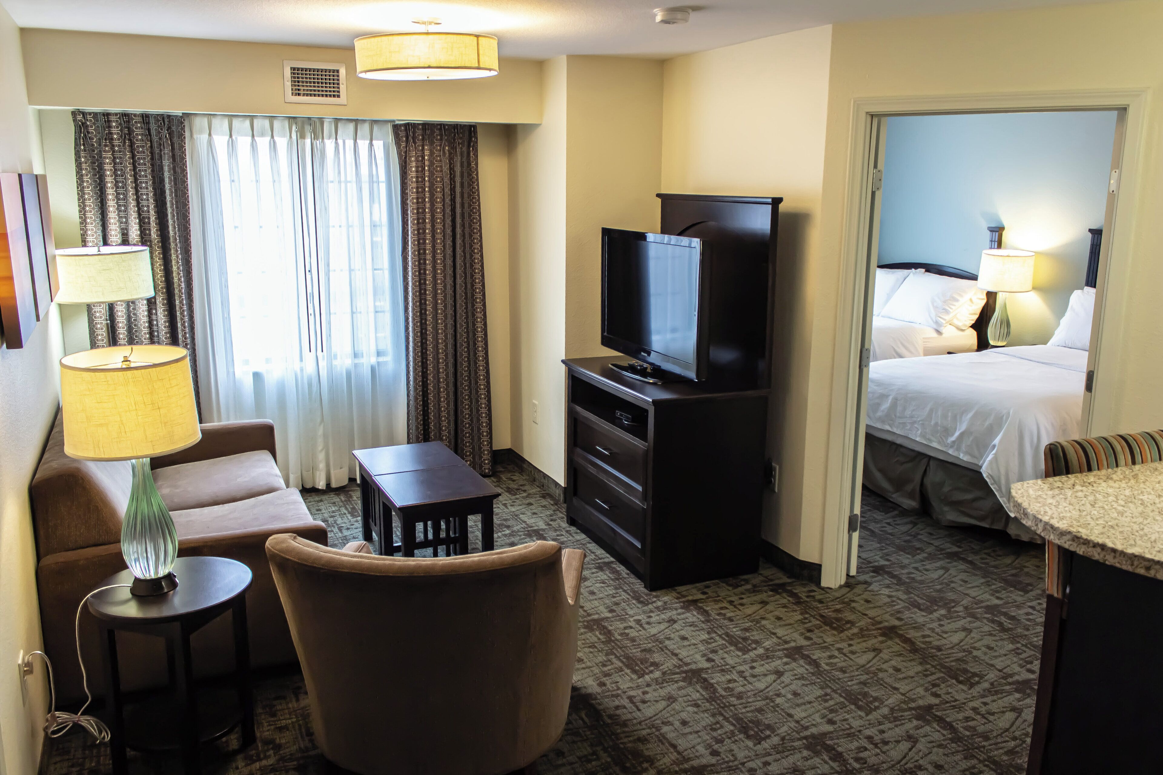 A hotel room with a sofa, a TV, a small table, a lamp, a bed, and a window with curtains.