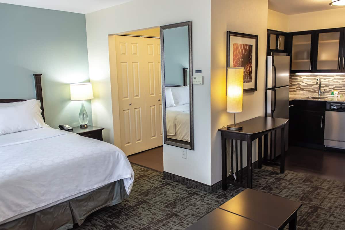 Una habitación de hotel con cama, mesita de noche, lámpara, espejo y una kitchenette con refrigerador y lavamanos.