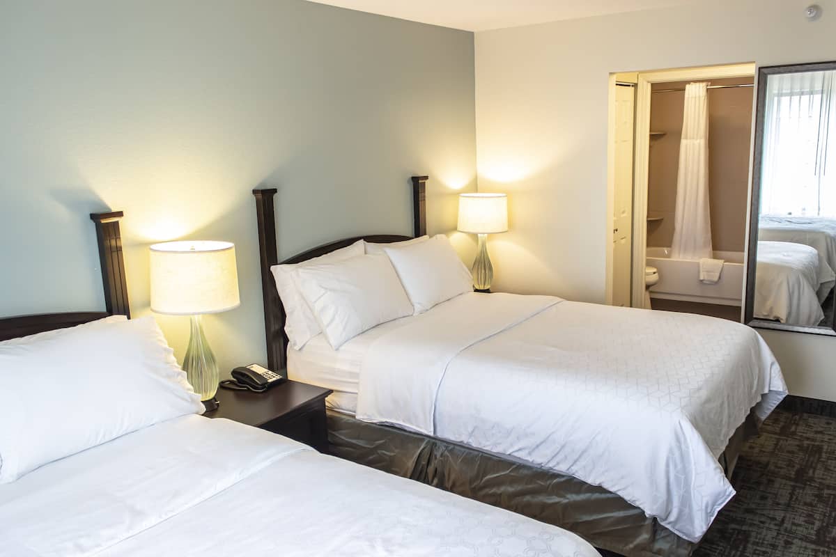 Habitación de hotel con dos camas, una mesita de noche con lámpara, teléfono y un espejo que refleja el baño.