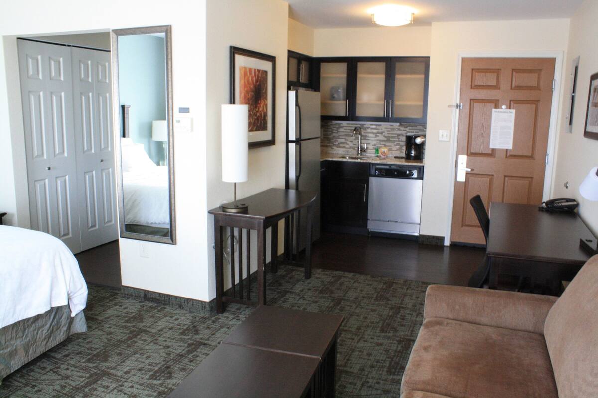 Una habitación de hotel con cama, un pequeño espacio de descanso con mesa y sofá, kitchenette y baño con ducha e inodoro.