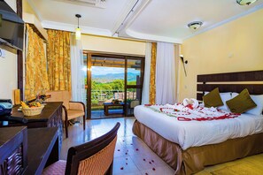 A hotel room with a bed, a desk, a chair, a TV, and a balcony with a view of the landscape. - Lake Bogoria Spa Resort (Bogoria)