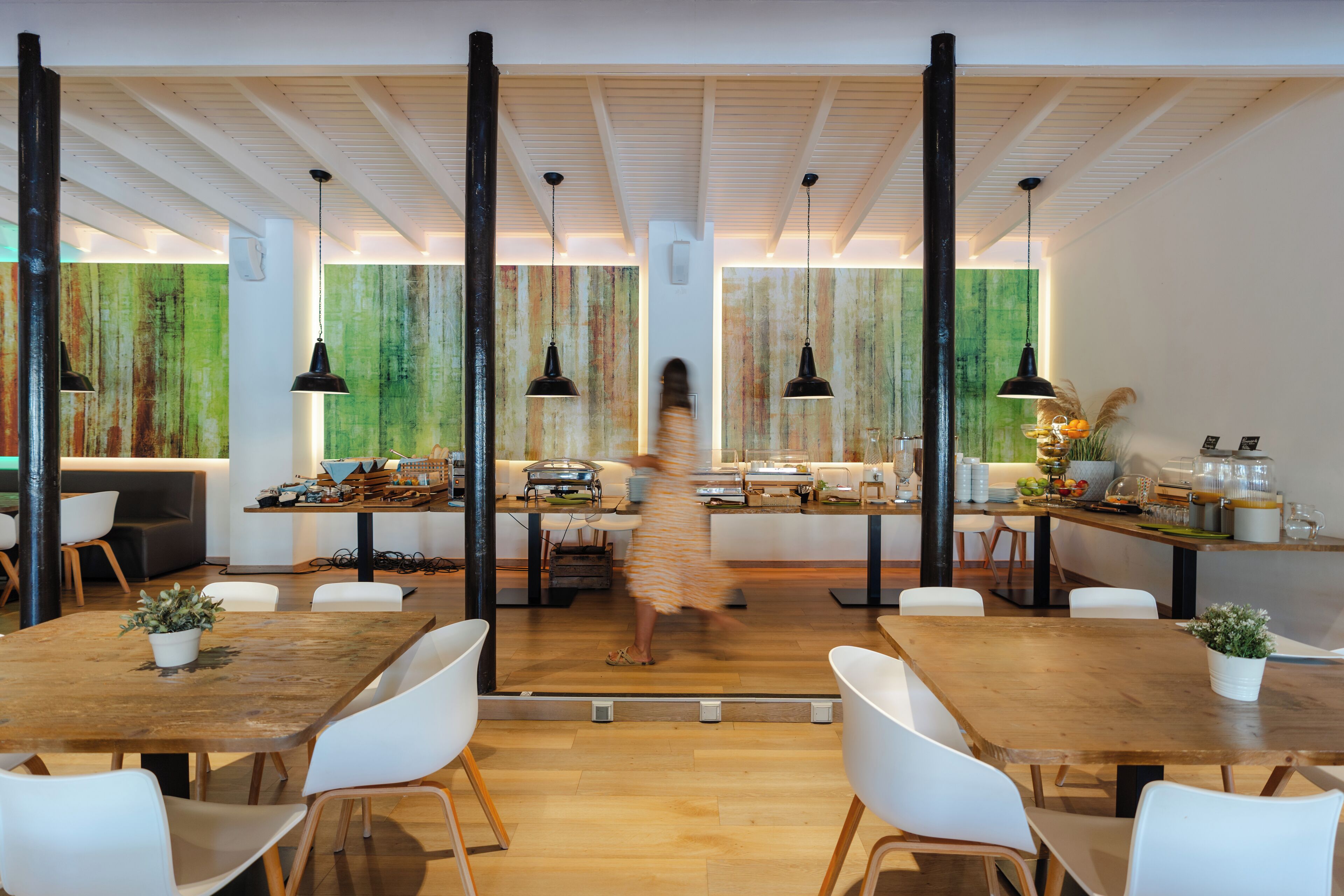 Un comedor moderno con mesas de madera y sillas blancas, que cuenta con un pasillo central e iluminación colgante.