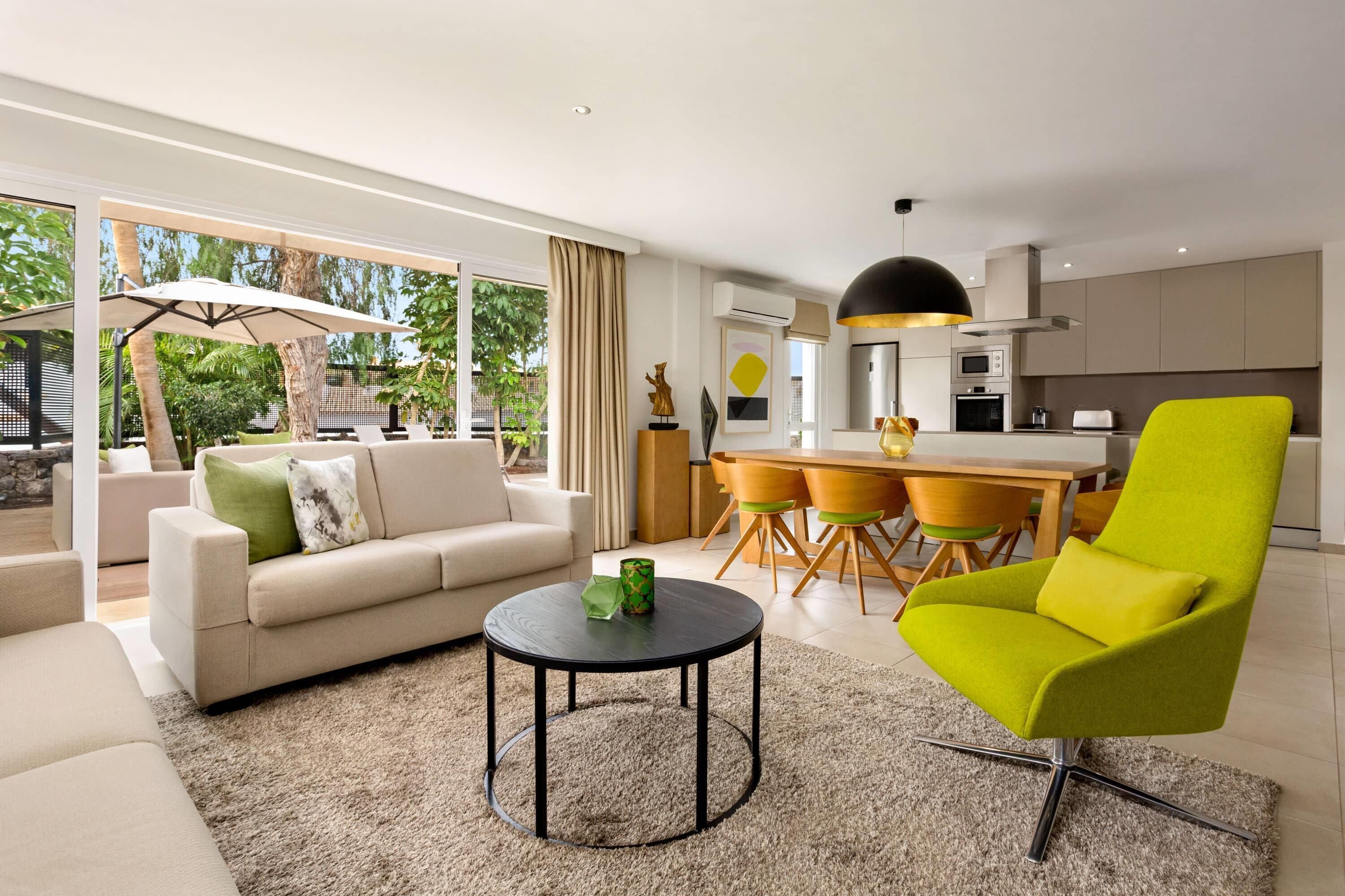 A modern living room with a sofa, a bright green chair, a round coffee table, and a dining area with a kitchen in the background.