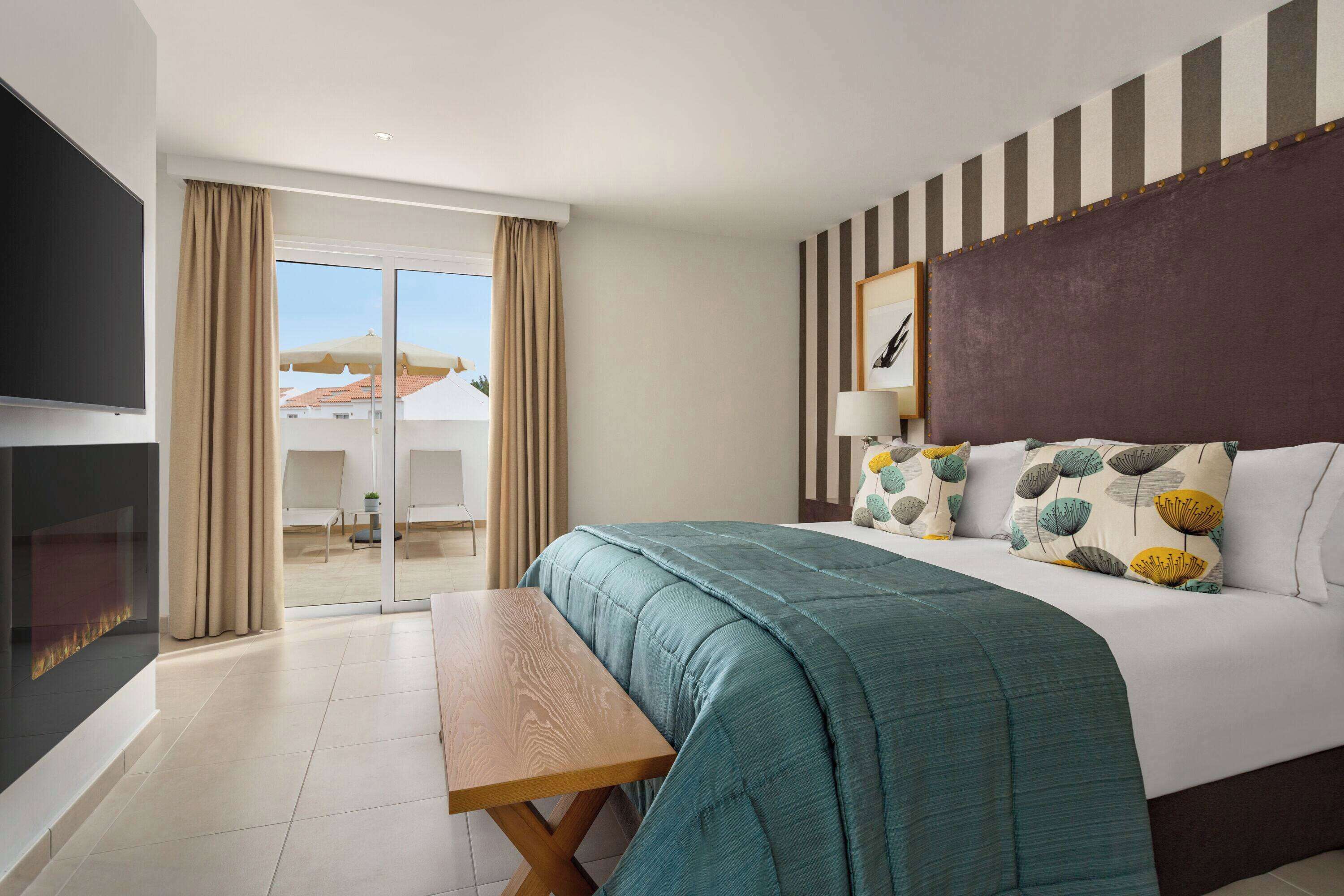 Habitación de hotel con una cama grande, un televisor de pantalla plana, un banco de madera y vistas a la ciudad desde la ventana.