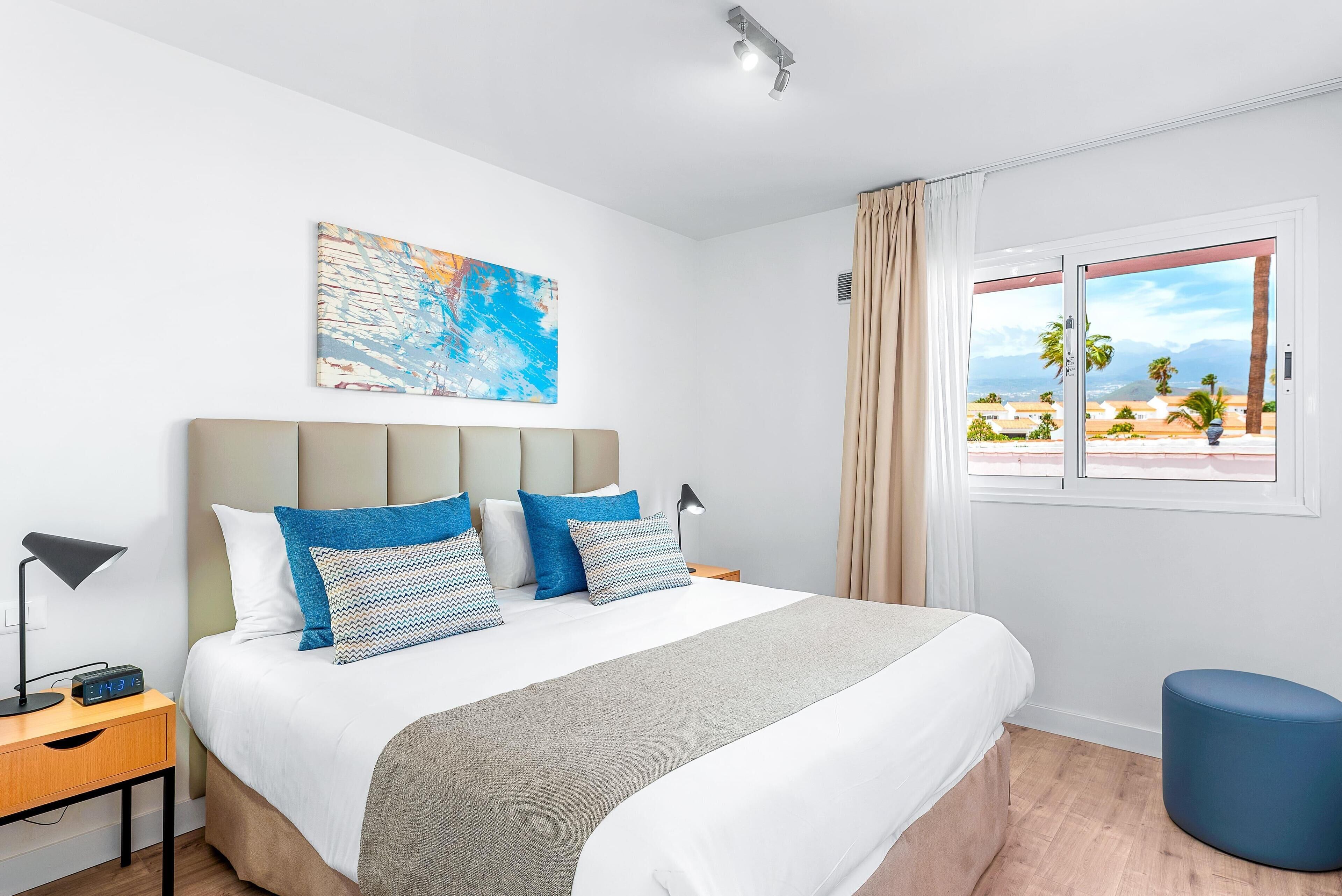 Een moderne hotelkamer met een groot bed, een nachtkastje met een lamp, een schilderij aan de muur en een raam met uitzicht op palmbomen en bergen.