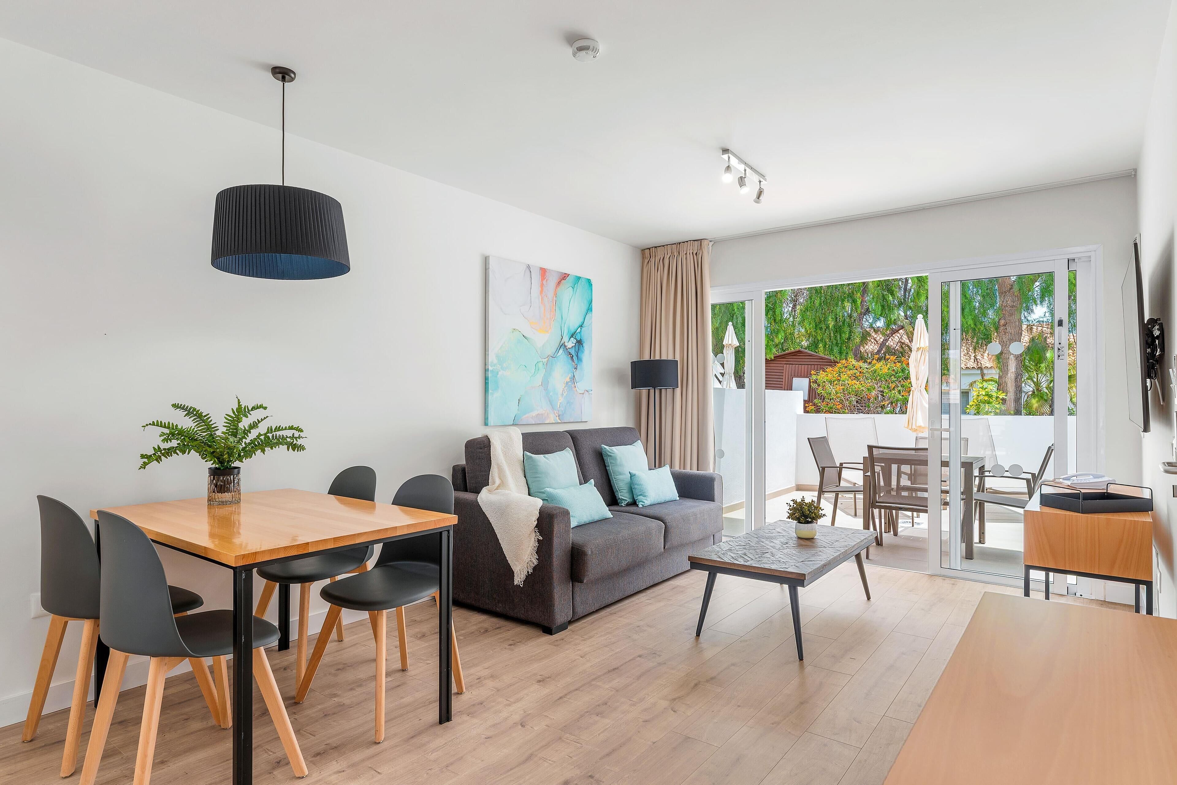 A modern living room with a sofa, dining table, and chairs. There is a large window with a view of greenery outside.