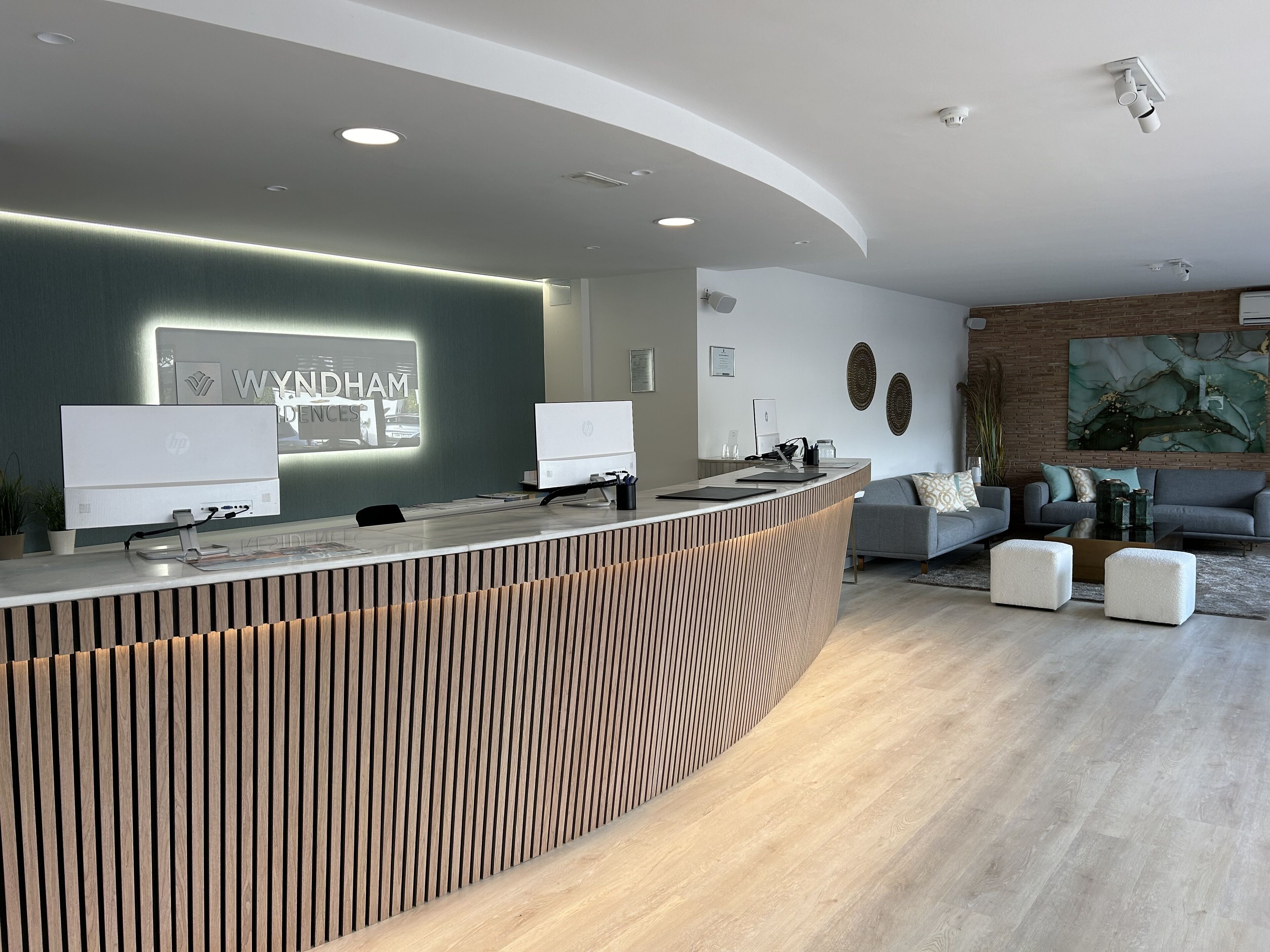 Un comptoir d’accueil moderne doté de deux écrans, d’un mur décoratif en bois et d’un espace salon avec un canapé gris et des poufs.