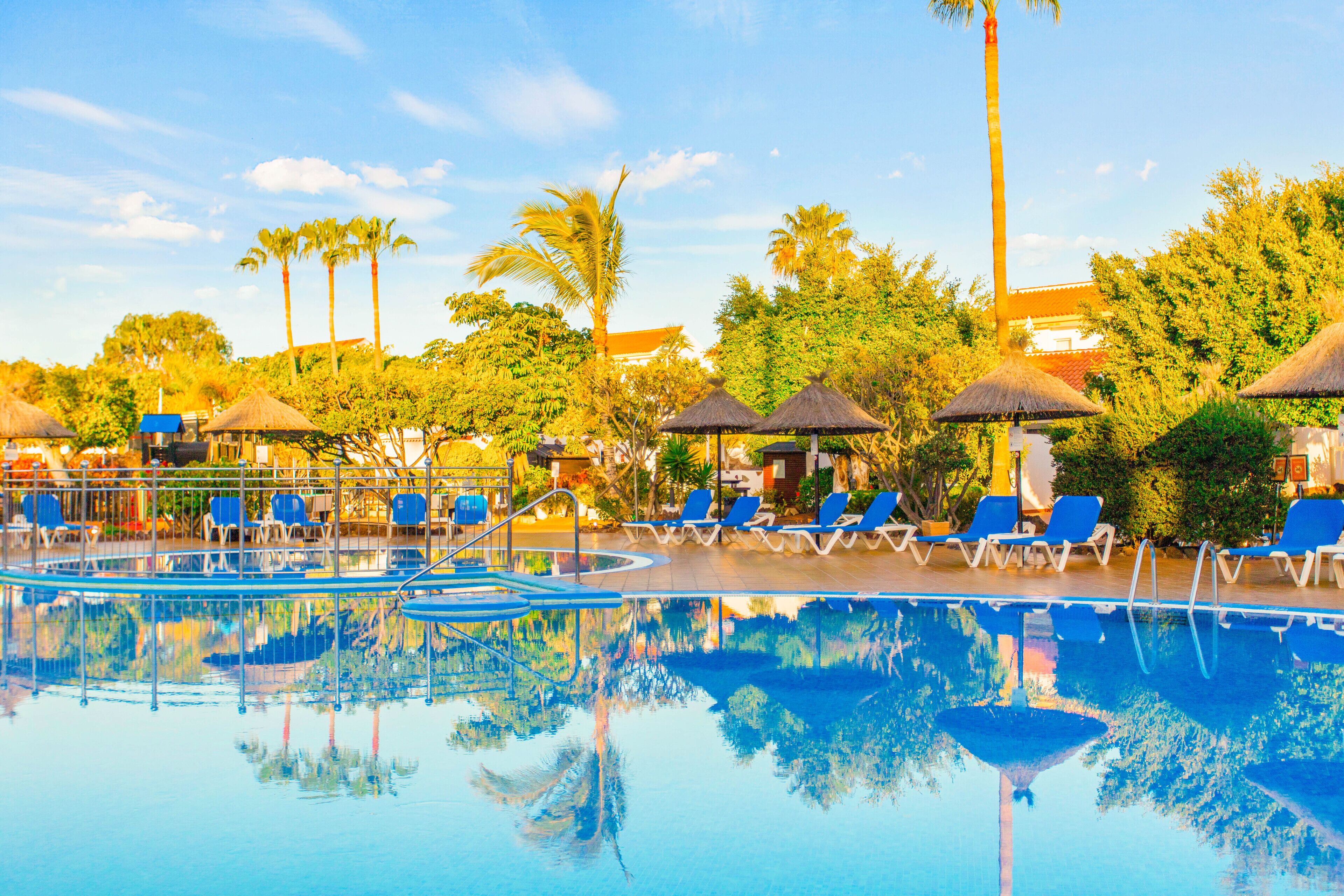 A pool area with lounge chairs and umbrellas, surrounded by palm trees and a building in the background.