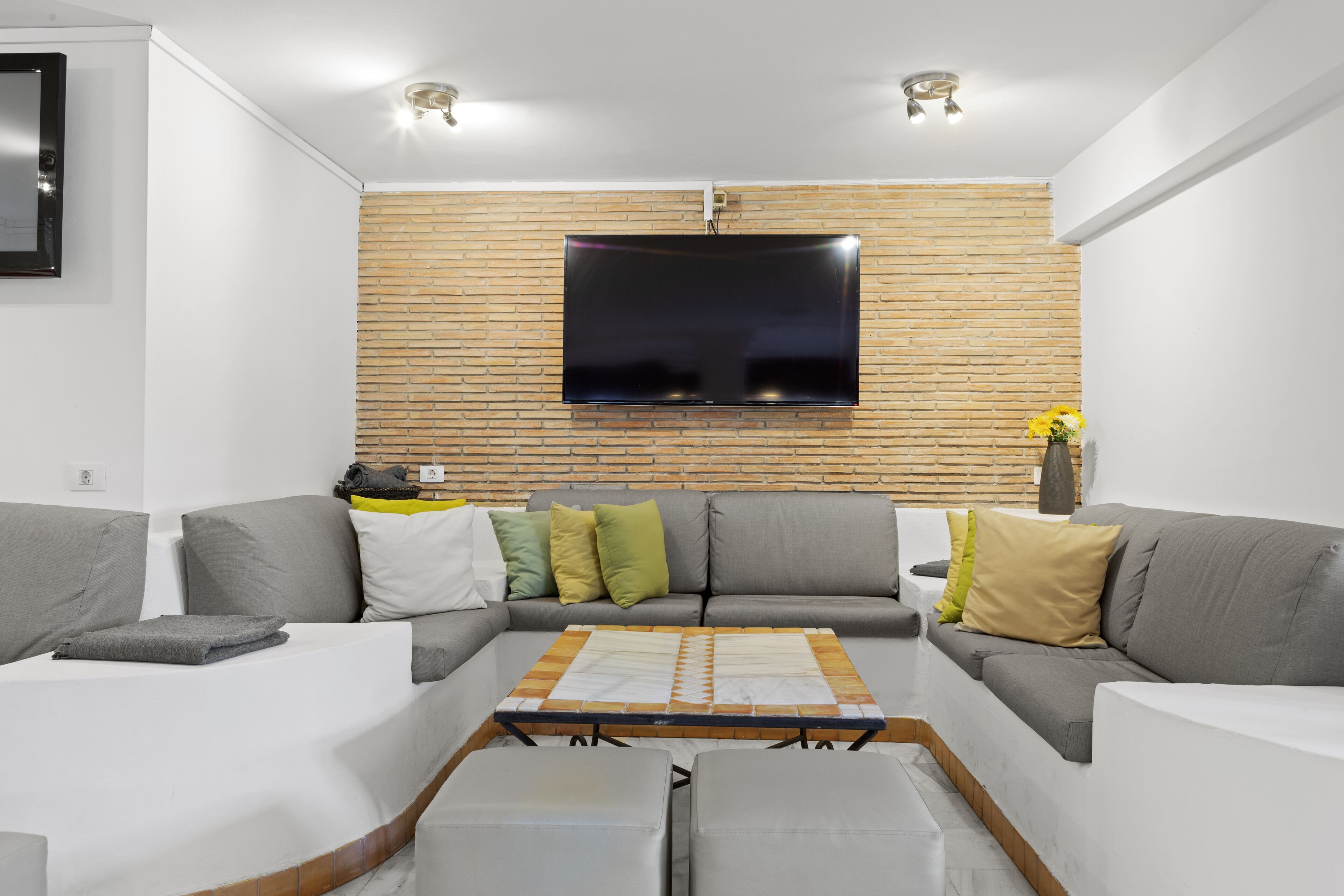 Un salon moderne avec un téléviseur à écran plat fixé à un mur en briques, des canapés gris avec des coussins, une table basse et une petite table d’appoint sur laquelle est posé un vase.