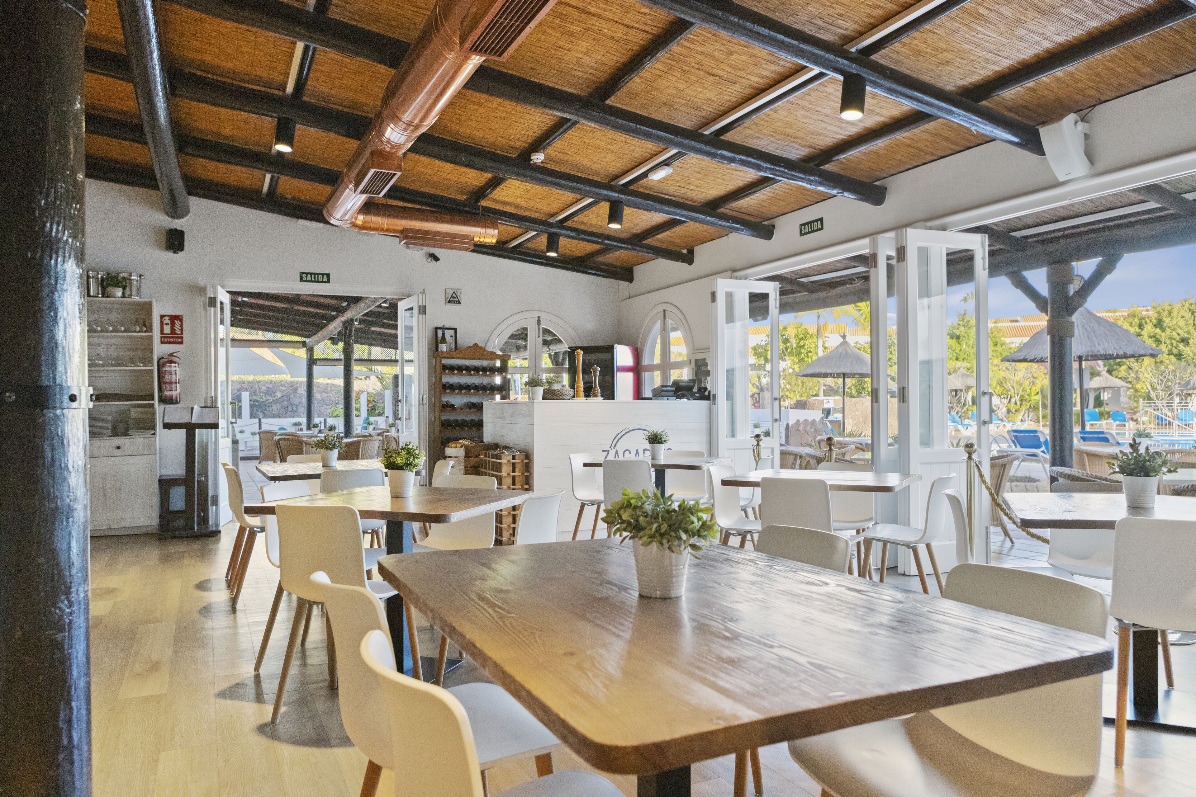 Un comedor amplio con mesas de madera y sillas blancas, una zona de cocina con armarios blancos y ventanas grandes que ofrecen vistas al exterior.