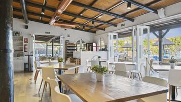Un comedor amplio con mesas de madera y sillas blancas, una zona de cocina con armarios blancos y ventanas grandes que ofrecen vistas al exterior.