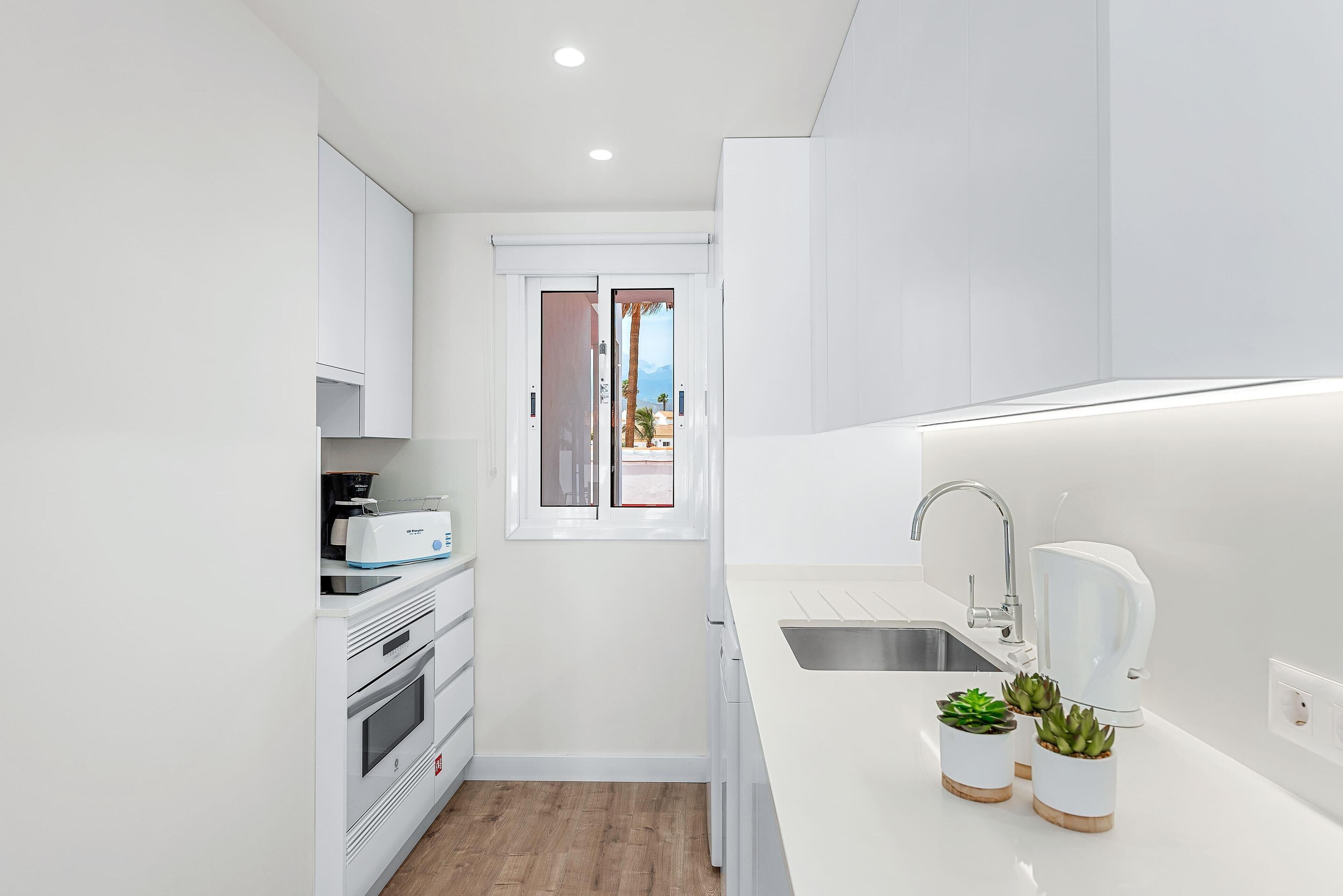 A modern kitchen with white cabinetry, a built-in oven, a microwave, and a sink with a faucet. There are three potted plants on the counter.