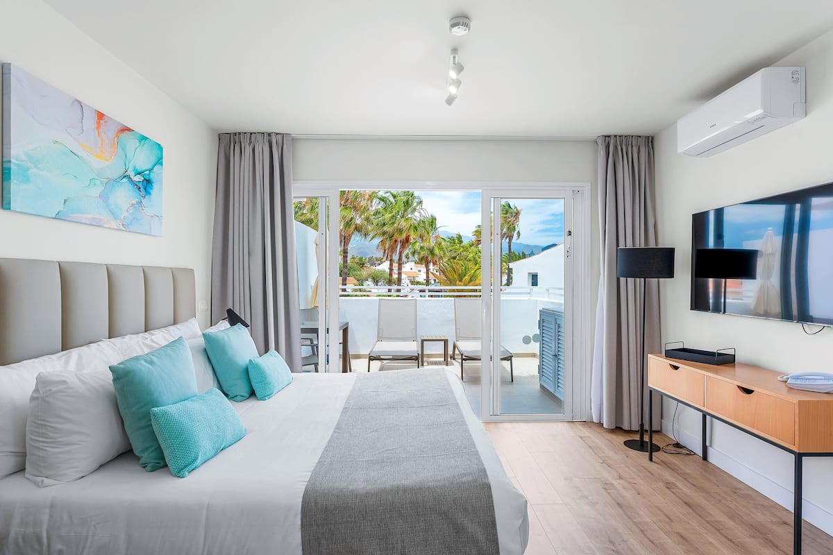 A modern hotel room with a large bed, a desk, a flat-screen TV, and a balcony with a view of palm trees and buildings.