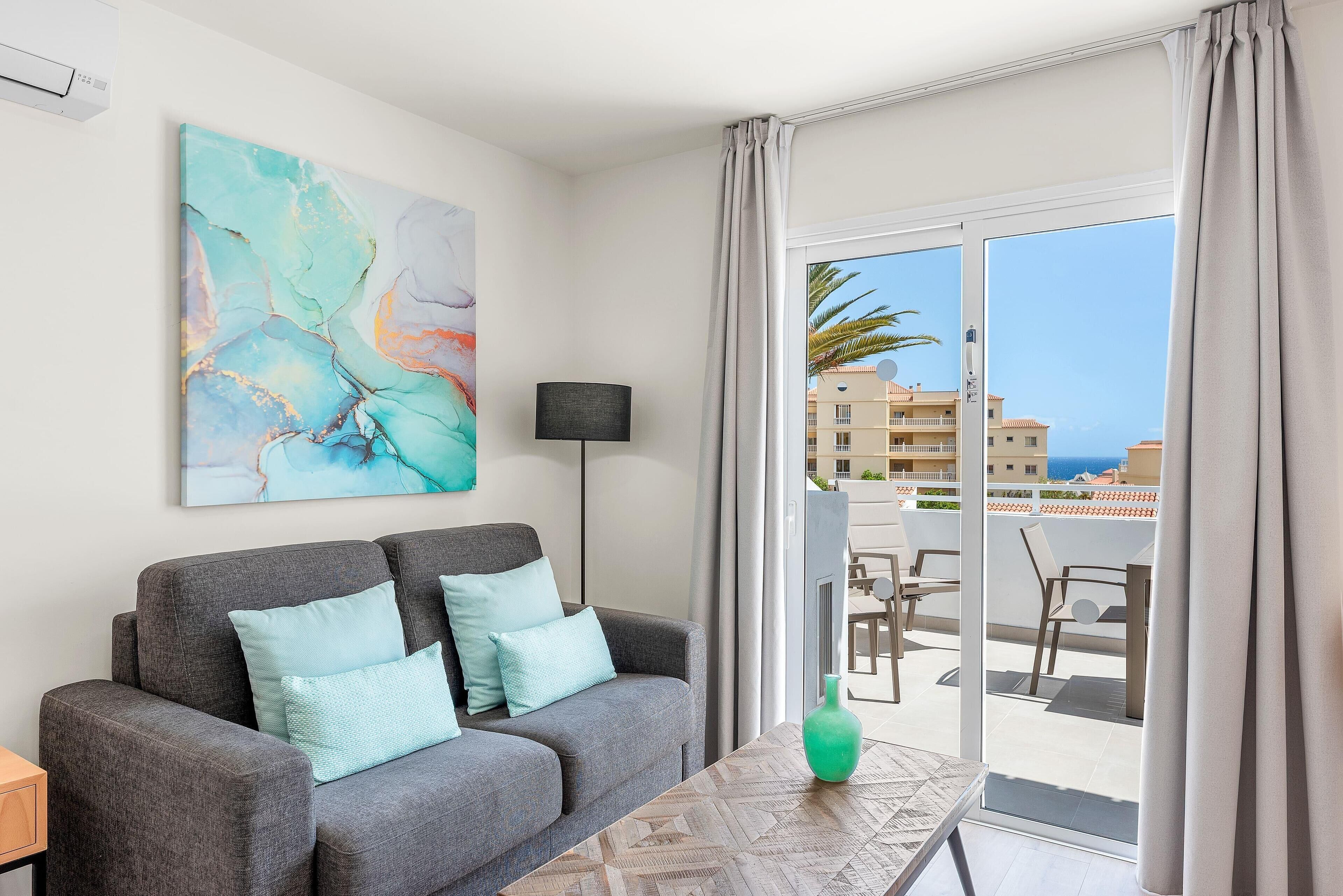 A modern living room with a grey sofa, teal cushions, a floor lamp, and a view of a balcony with outdoor furniture.