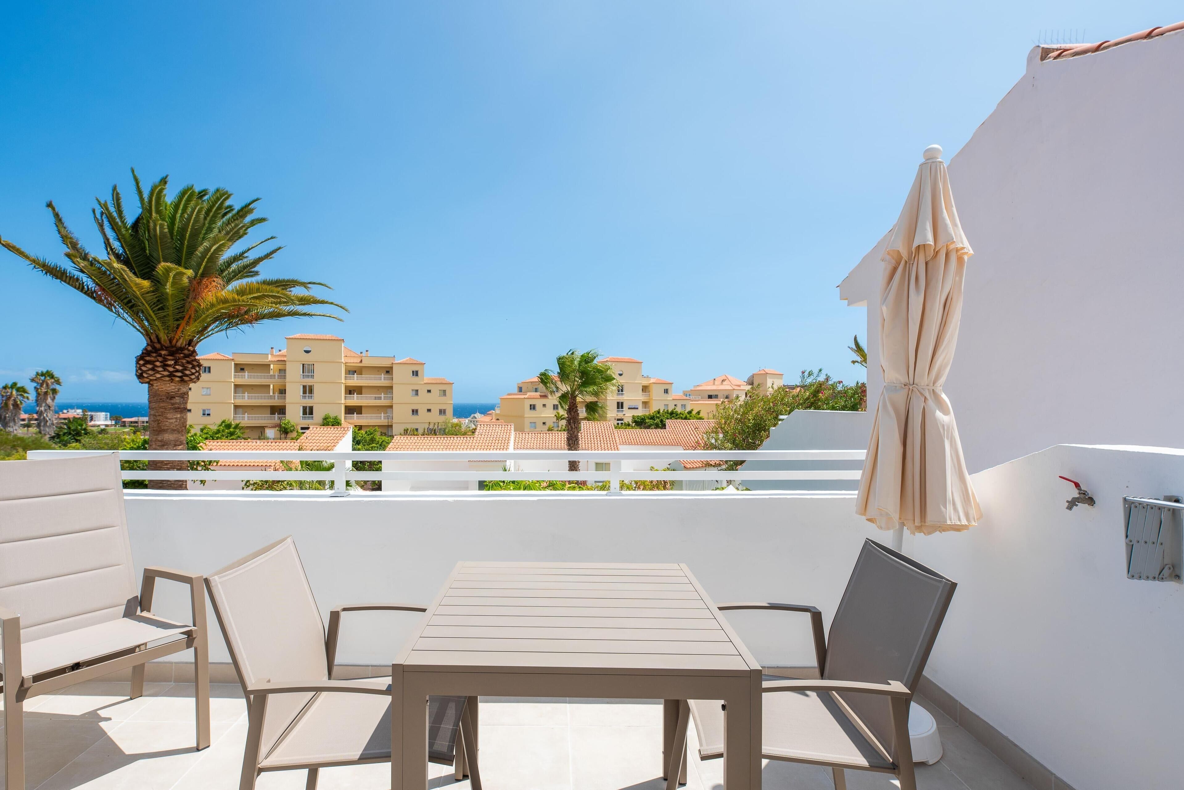 A balcony with a table and chairs, a palm tree, and a view of buildings and the sea.