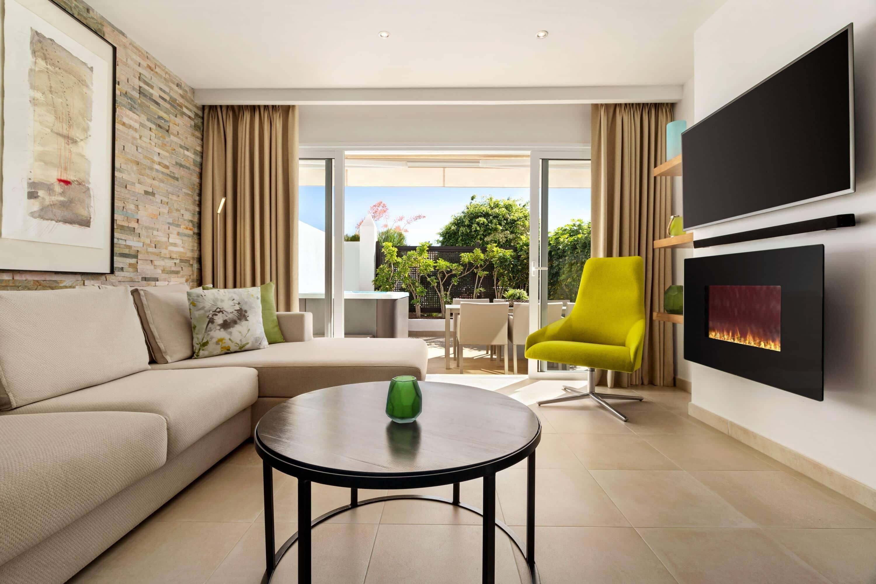 A modern living room with a sofa, a round table with a green vase, a bright yellow chair, a fireplace, and a large TV.
