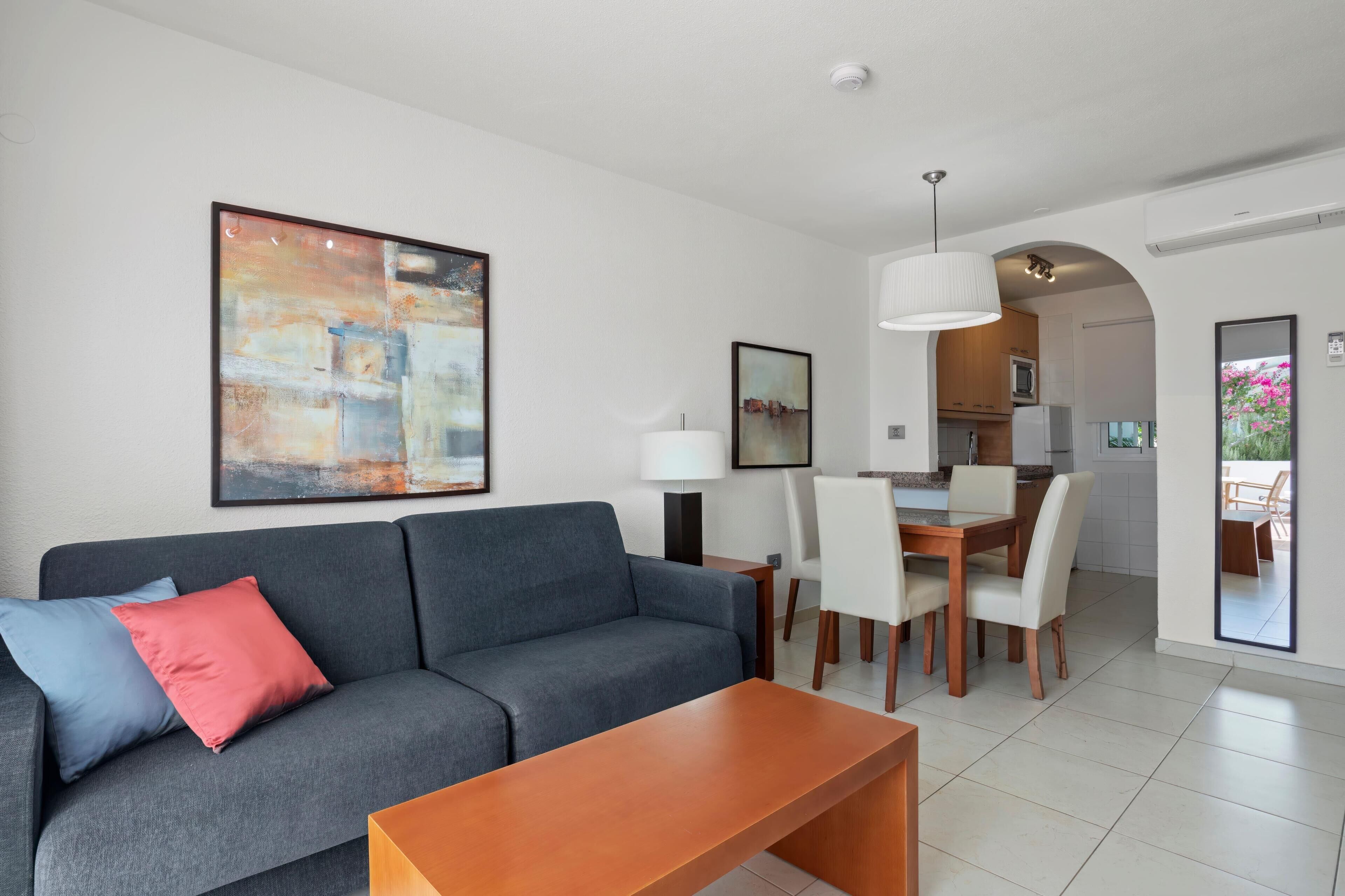 Una sala de estar moderna con un sofá gris, una mesa de centro de madera y un comedor con sillas blancas y una mesa de madera.
