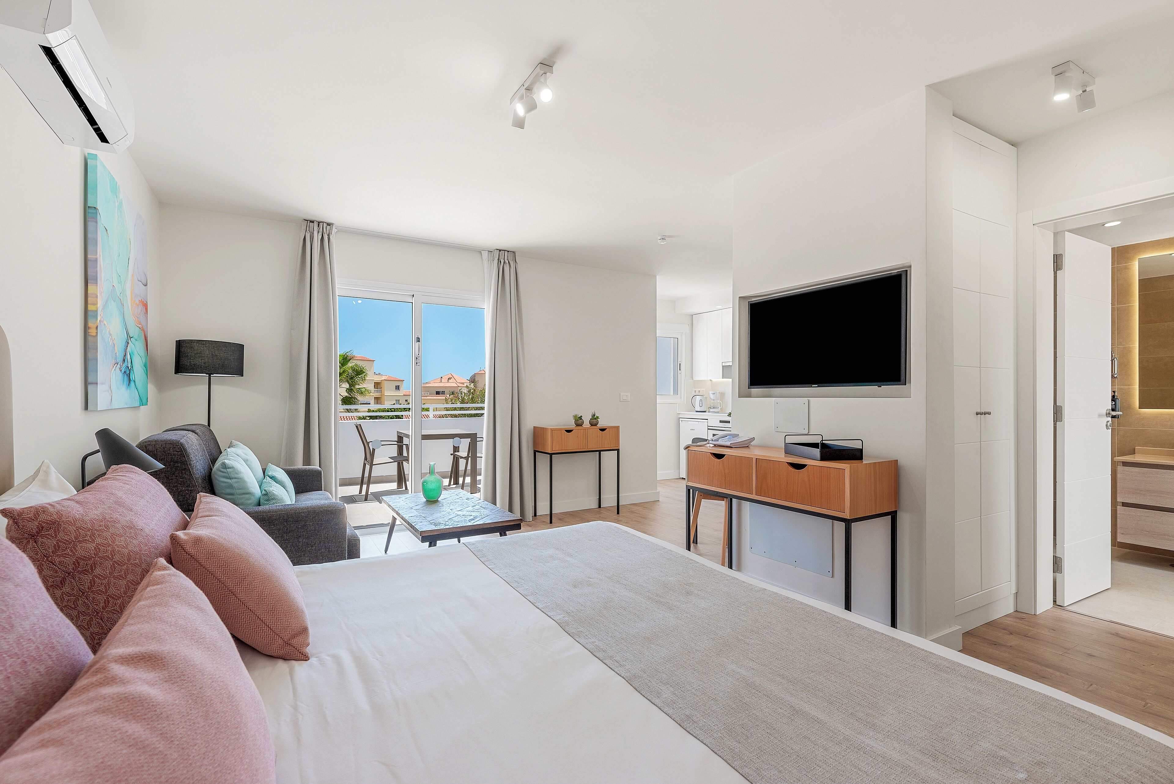 A modern hotel room with a large bed, a desk, a flat-screen TV, and a balcony with a view of the city.