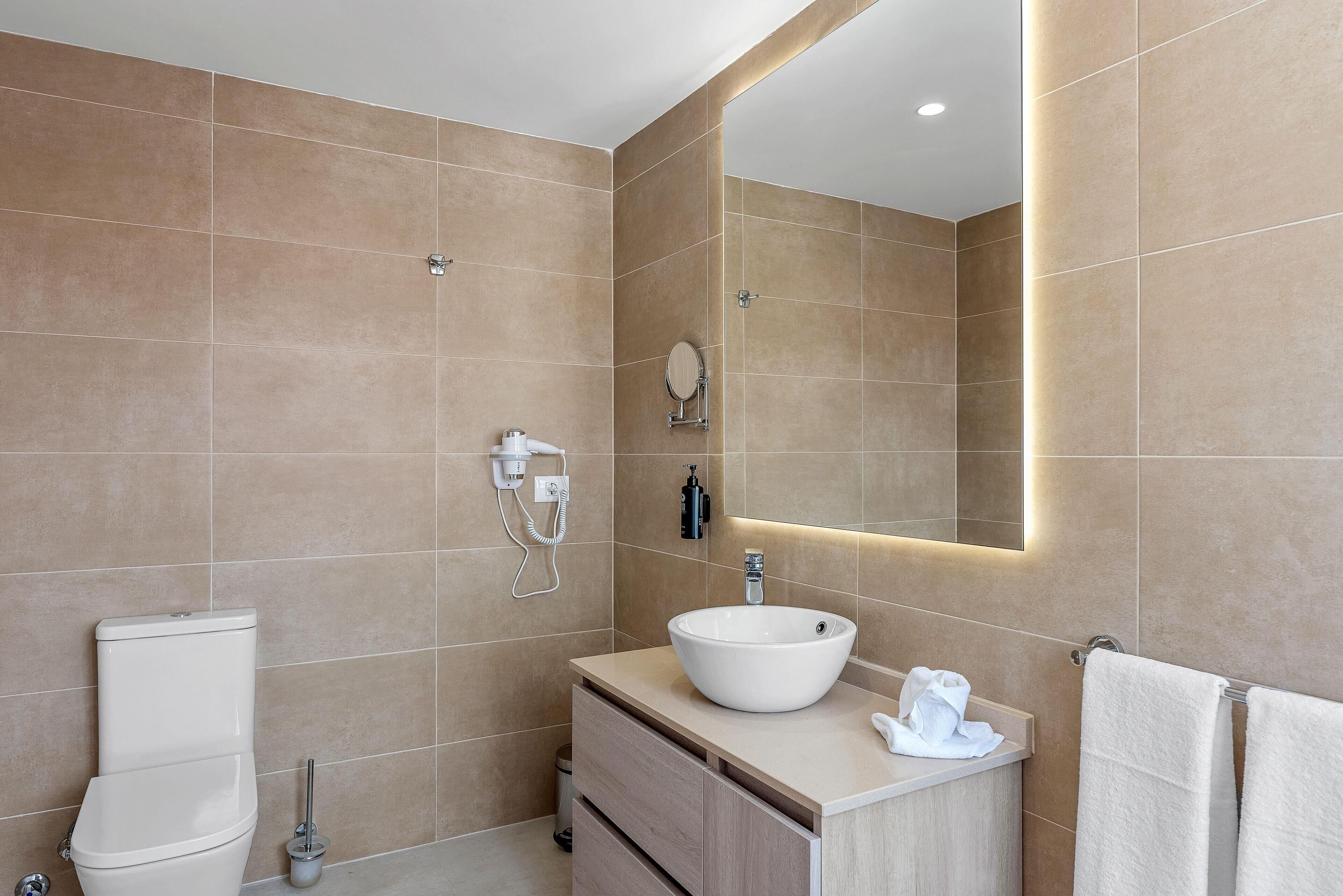 A modern bathroom with beige tiles, a white sink, a toilet, a vanity with drawers, a mirror, and a towel rack.