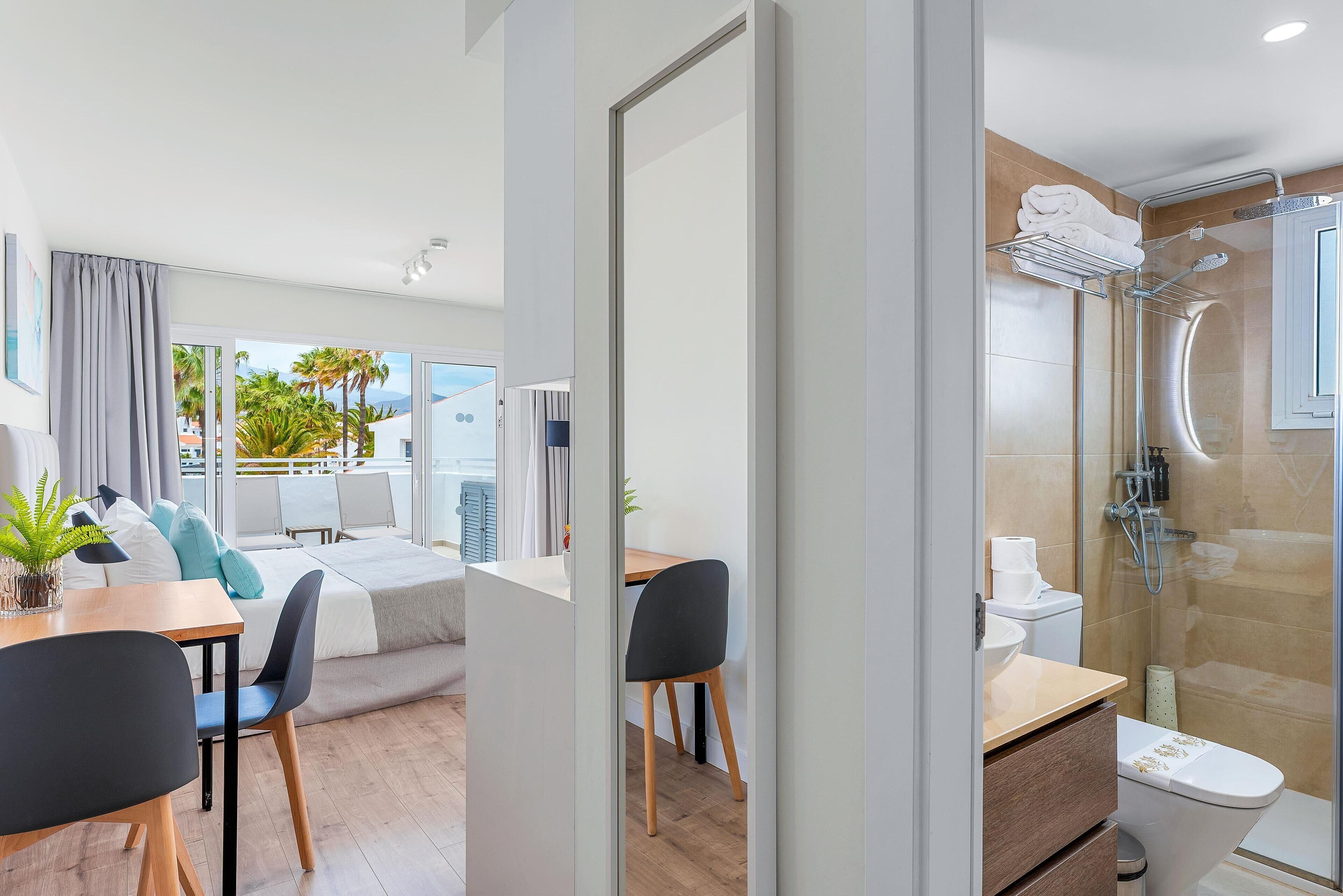 A modern hotel room with a balcony, a shower area, and a dining area with a table and chairs.