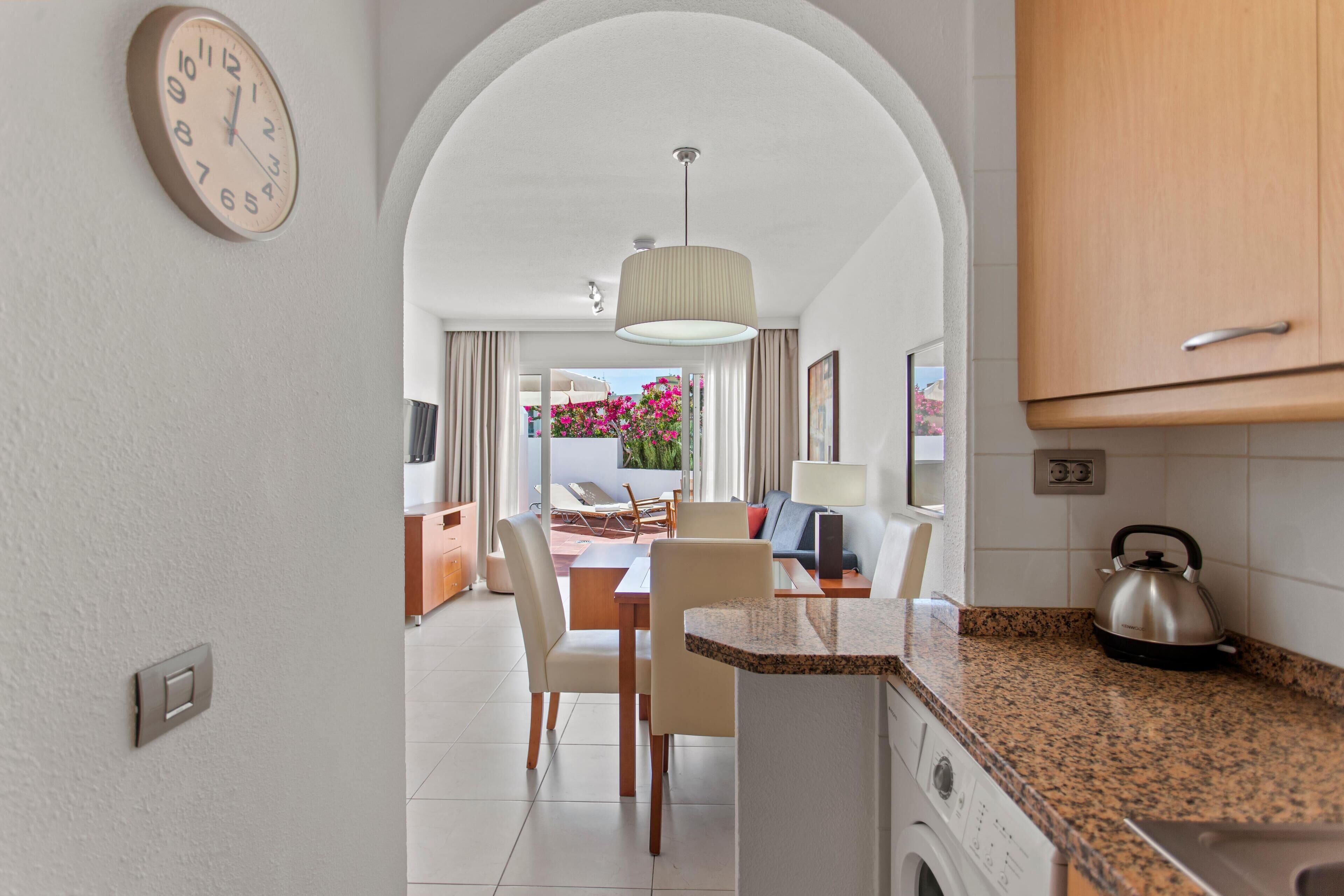 A modern kitchen with granite countertops, a kettle, and a dining area with a table and chairs.