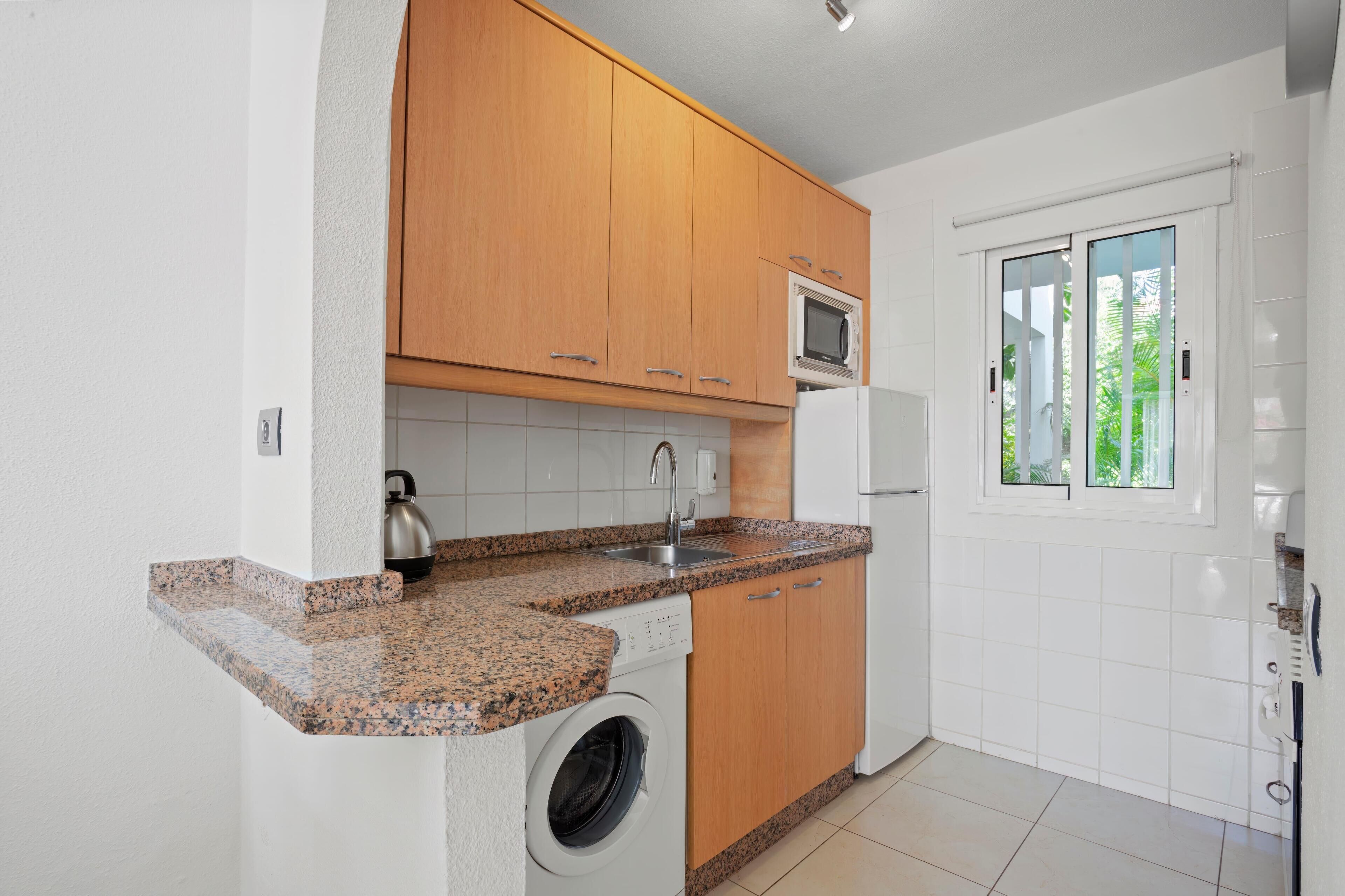 A compact kitchen with wooden cabinets, a built-in microwave, a washing machine, and a window with a view of greenery.