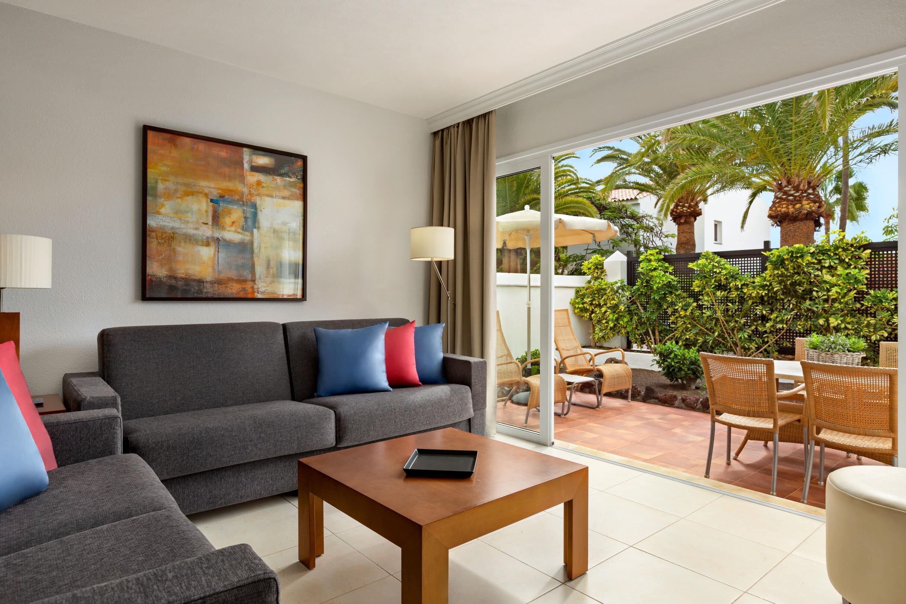 Un salón moderno con un sofá gris, una mesa de centro de madera y un amplio ventanal con vistas a un patio con muebles de exterior y vegetación.