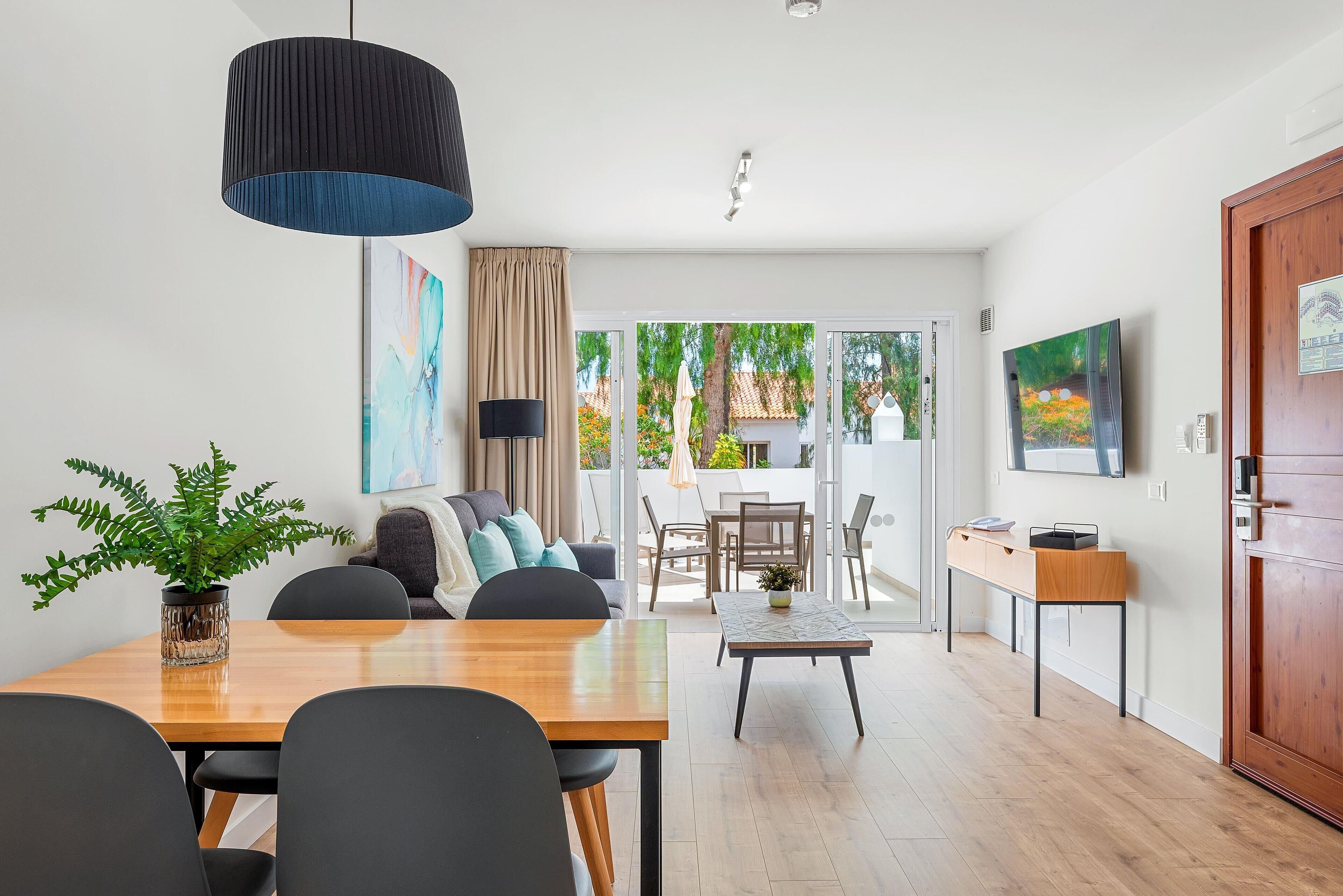 Una sala de estar moderna con una mesa de comedor de madera, sillas negras, una mesa de centro y una planta grande en maceta.