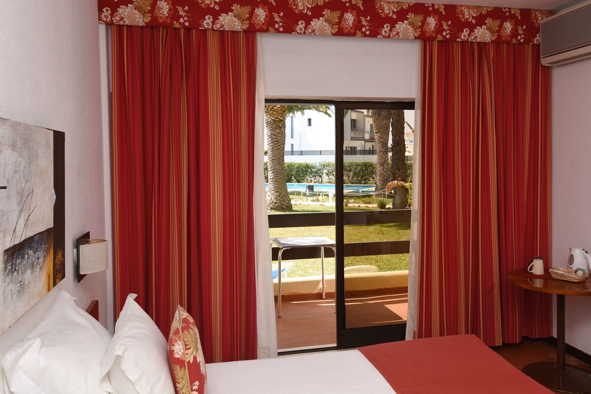 A hotel room with a bed, a table with a kettle, and a view of a pool and outdoor seating.