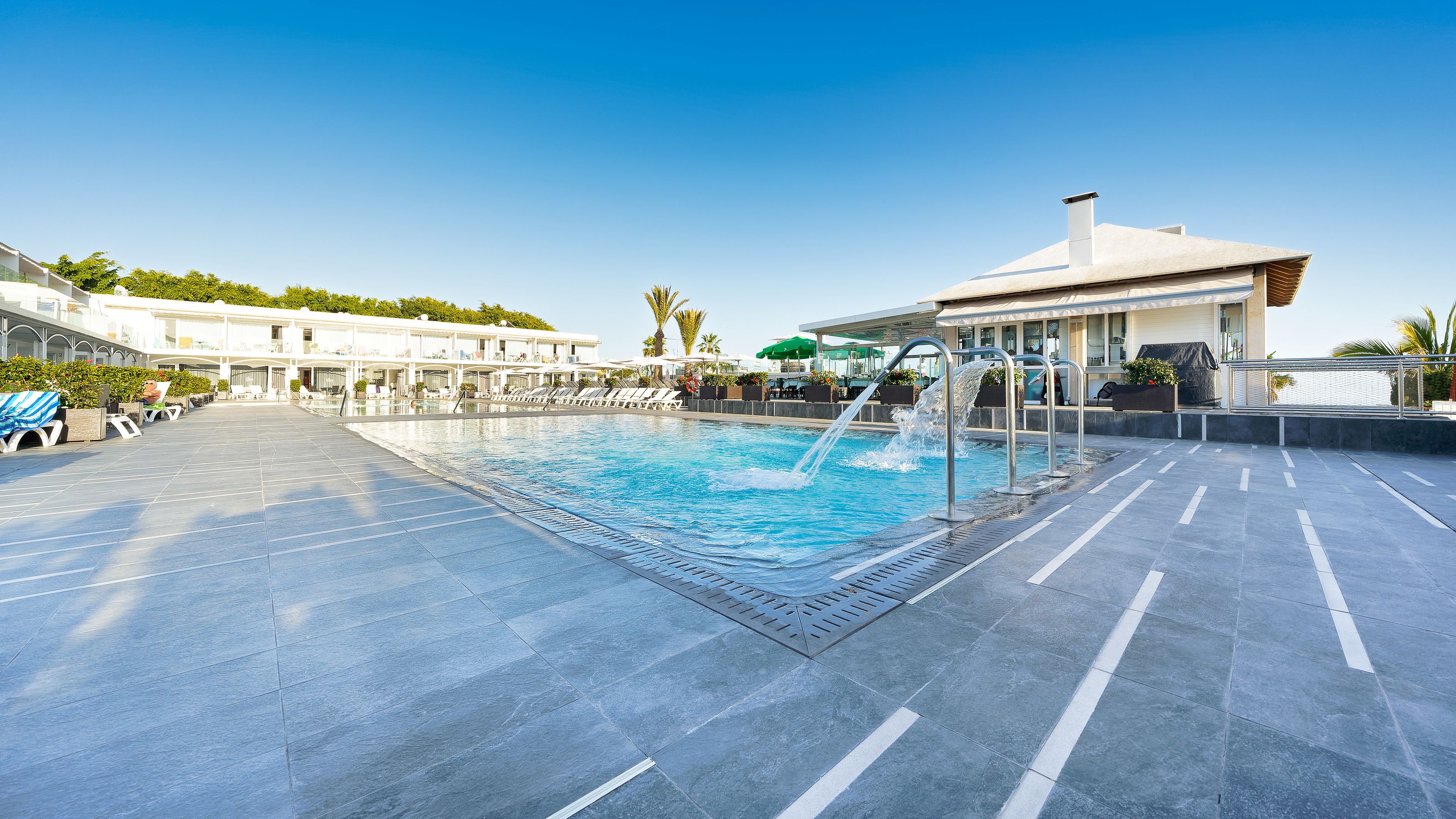 A rooftop pool with a water feature, lounge chairs, and a building with a white facade.