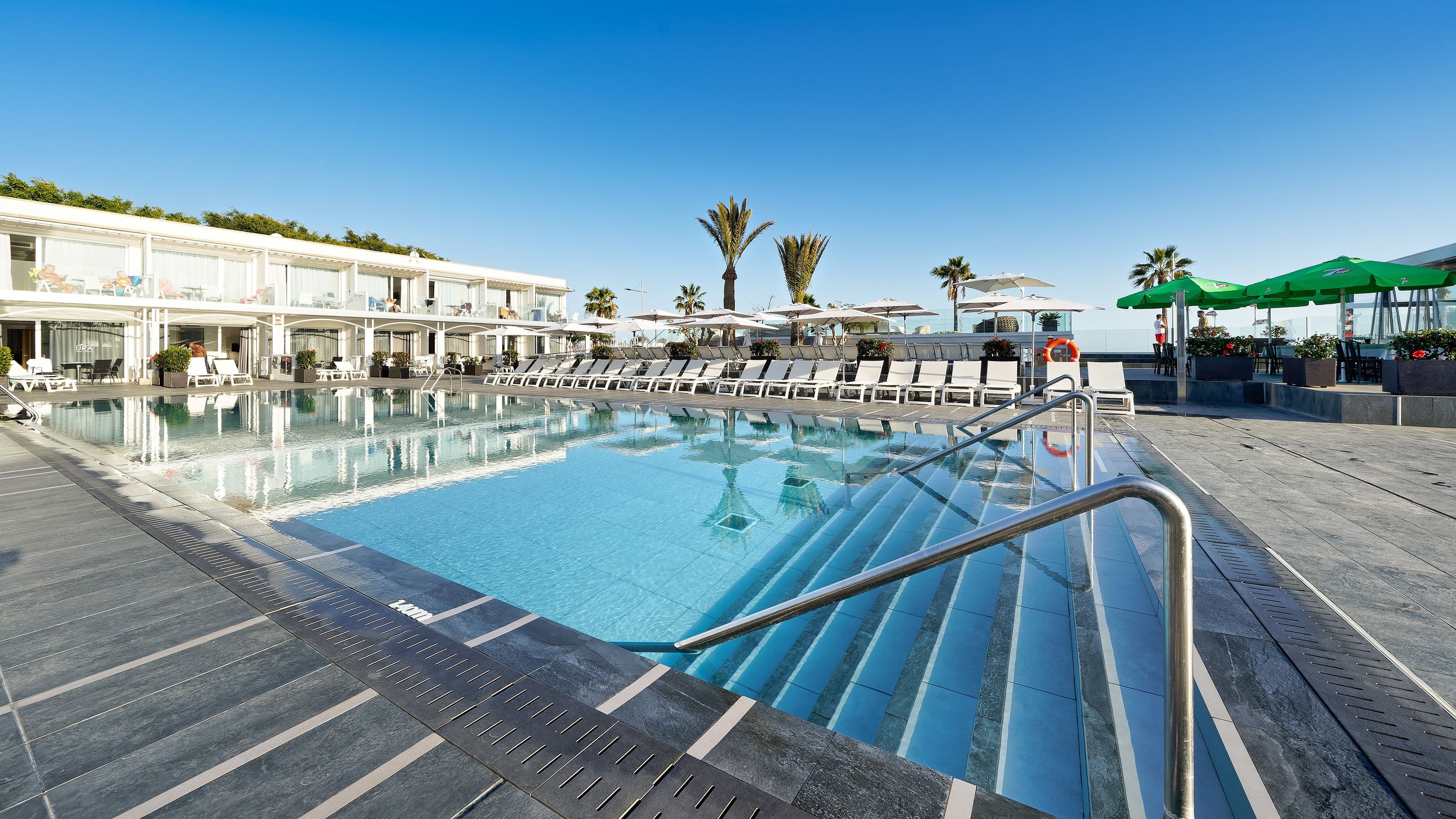 Una piscina con sillones de descanso y sombrillas, rodeada por un edificio de fachada blanca y un cielo azul despejado.