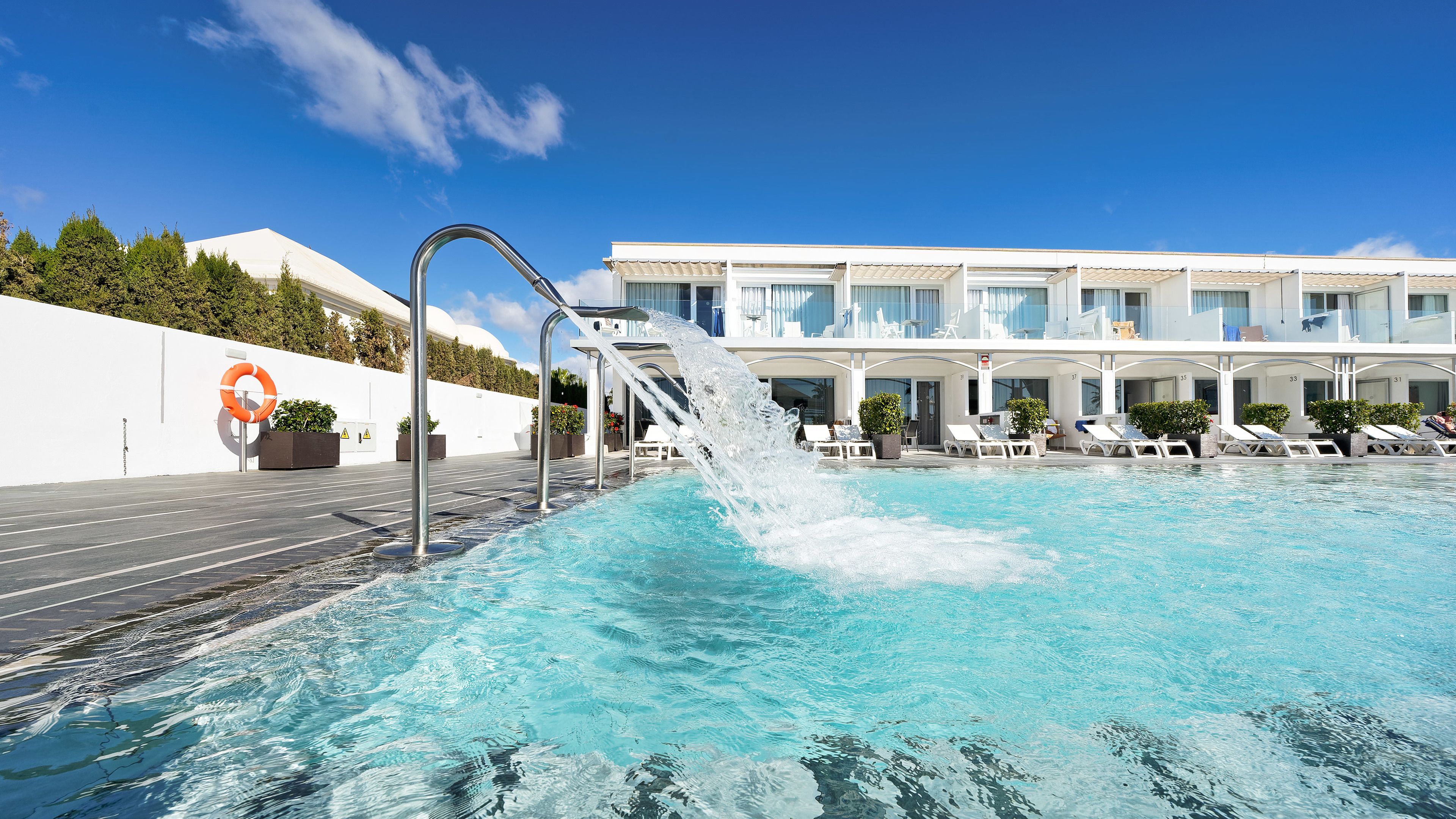 A swimming pool with a water feature, lounge chairs, and a building with numbered rooms in the background.