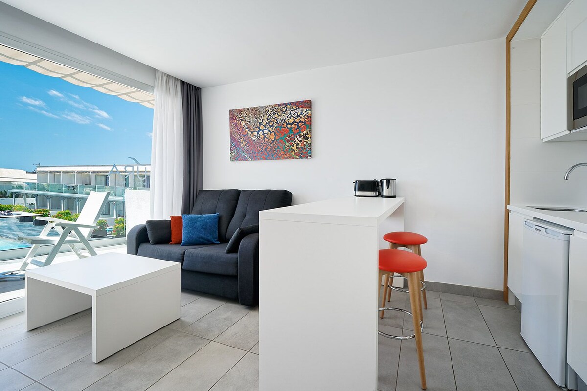 Una sala de estar moderna y bien iluminada con un sofá, una mesa de centro y una cocina con microondas y lavadero.
