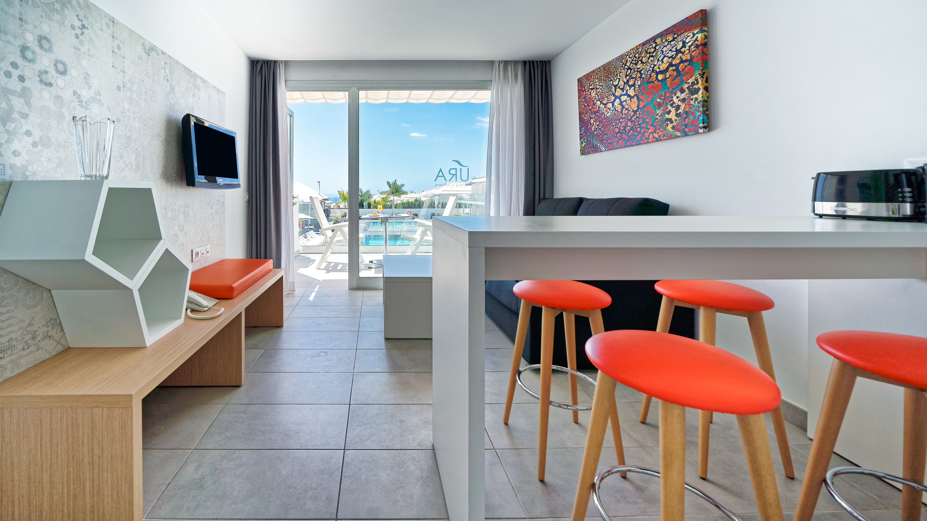 Une chambre d’hôtel moderne avec vue sur la piscine, un bureau avec un ordinateur portable, une télévision et une peinture abstraite aux couleurs vives.