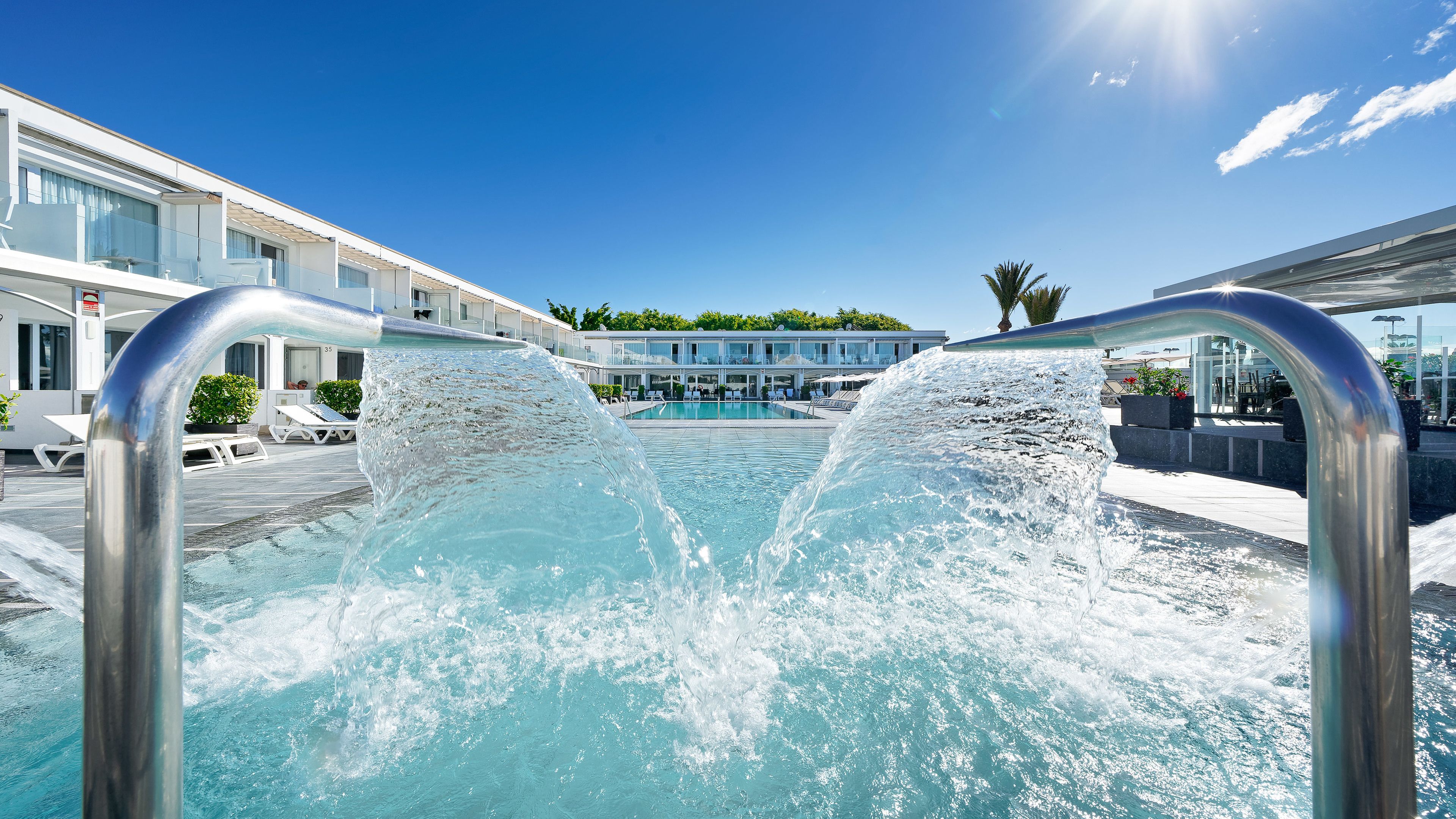 Una piscina con cascada, rodeada de sillas de descanso y un edificio con amplias ventanas.
