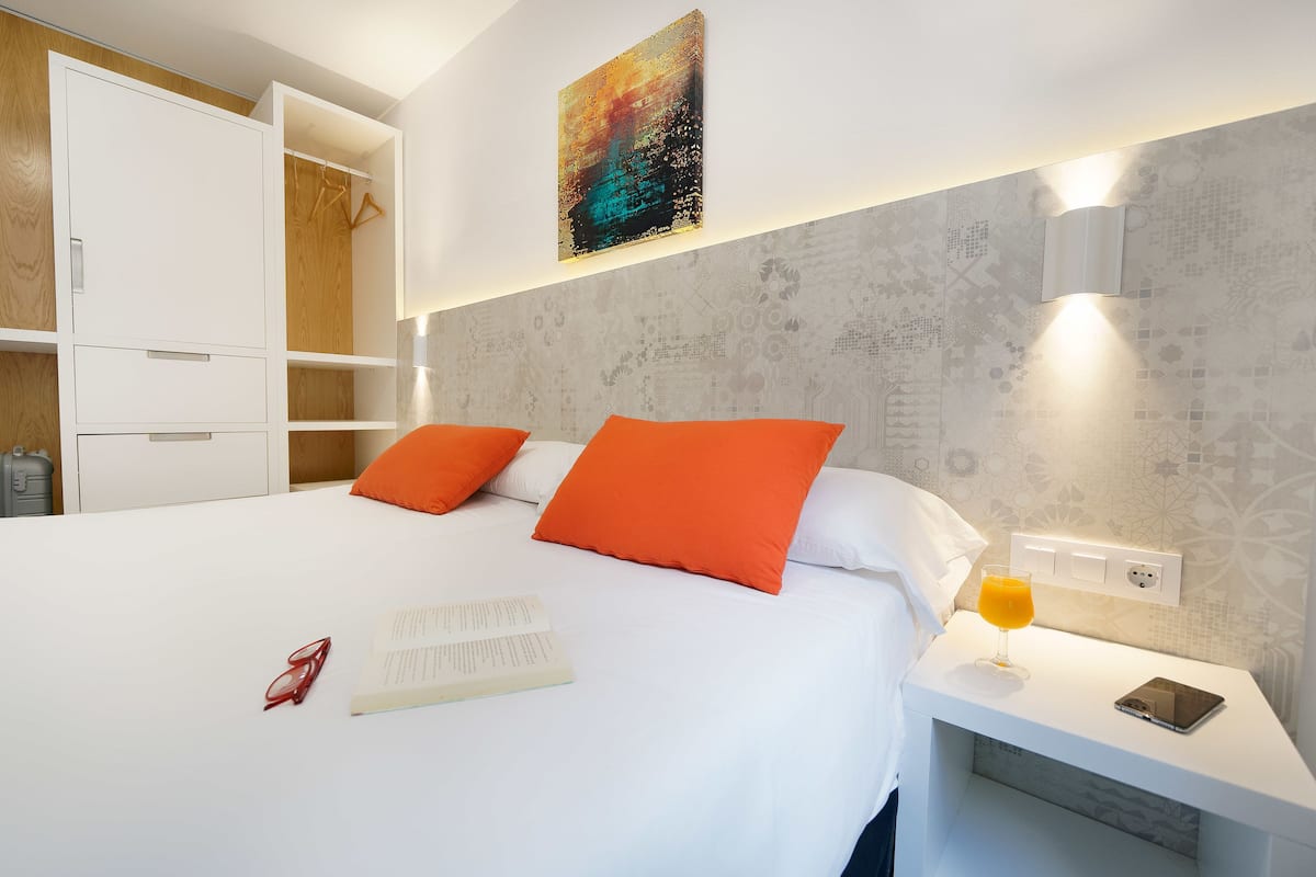 A hotel room with a bed, two orange pillows, a bedside table with a glass of orange juice, a book, and a pair of glasses.